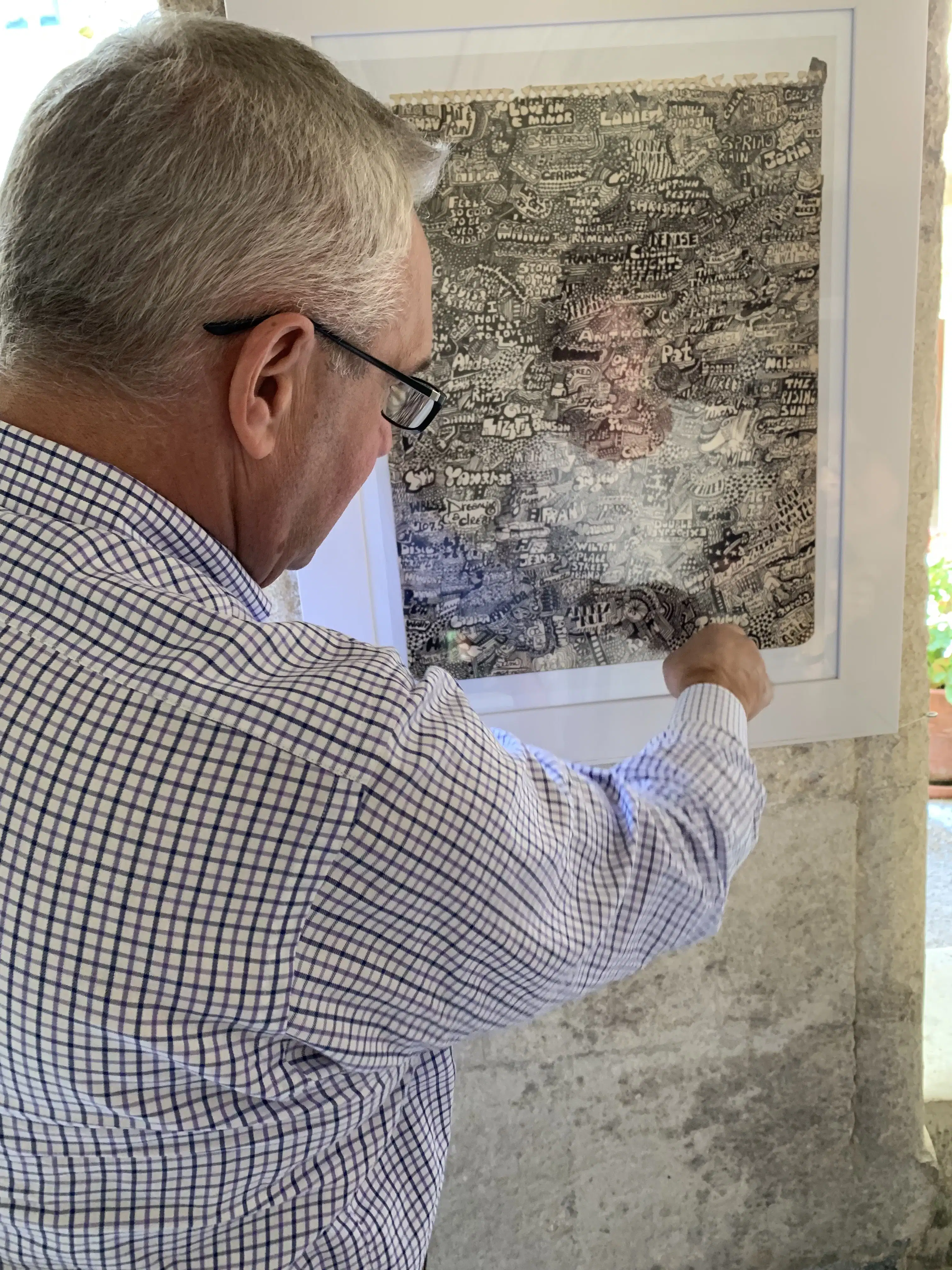 A man points to a detail in a framed print on the wall. The print is a page filled with words and shapes in black ink.