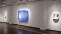 Exhibition photograph of “Between Picture and Viewer," showing three paintings on a wall. In the center is a large abstract in blue, white, and orange, and on either side of it is a single painting in white with red markings.