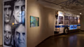 To the left is an oversized print of four portraits in black and white, to the right is a monitor playing a video and a cut-in-half New York City MTA bus protruding from wall.
