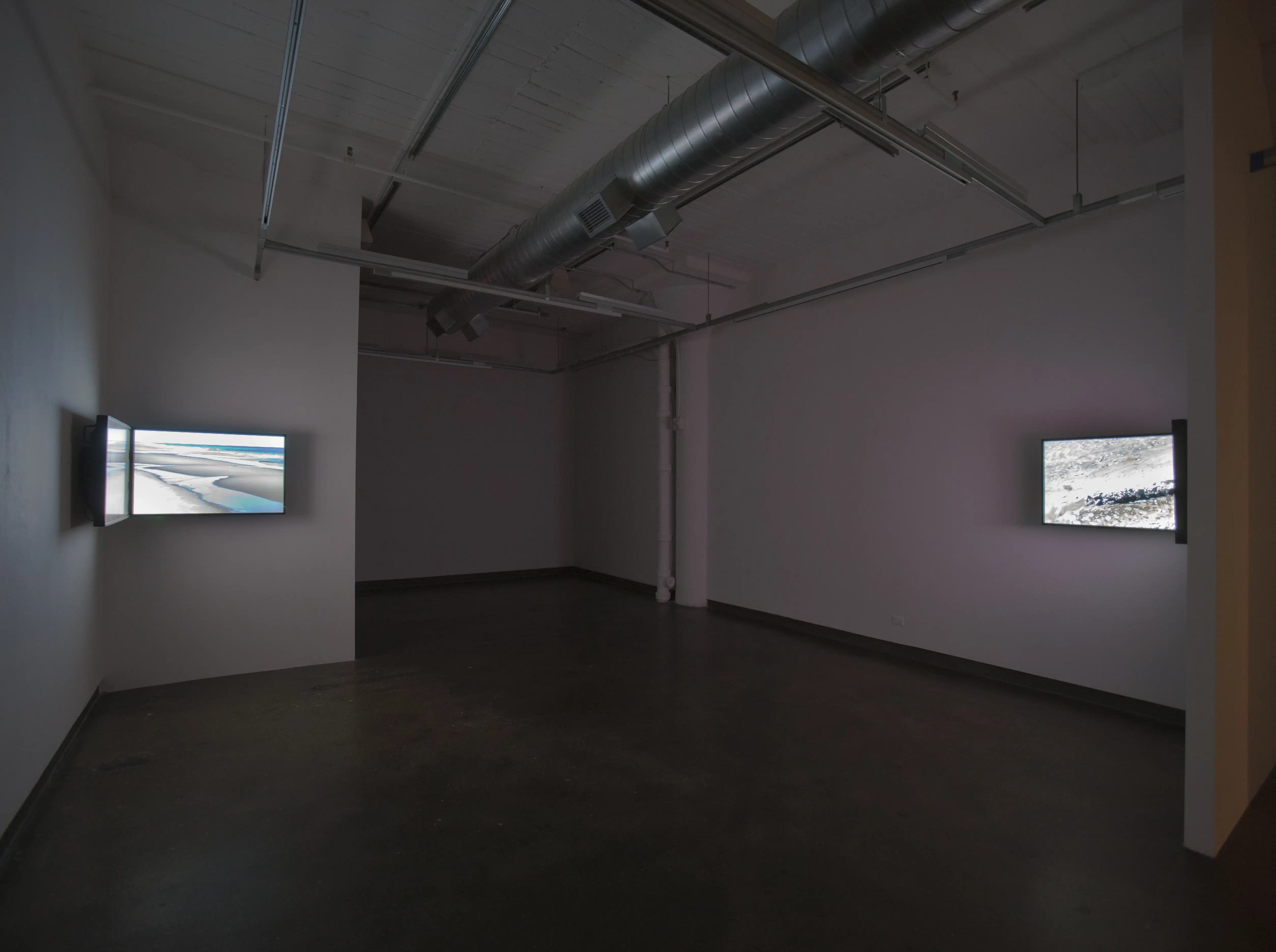 Dark room with lit photograph boxes in opposing corners. The ceiling is not finished, and there are pipes running along it.