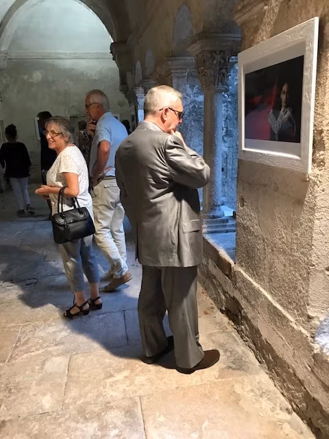 A man in a gray suit looks closely at a framed photograph on the wall. More visitors stand in the hall behind him.