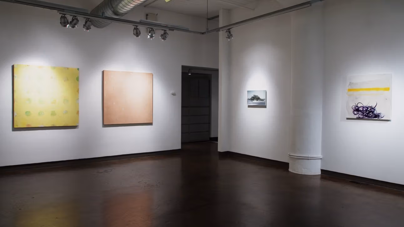 Exhibition photograph of “Between Picture and Viewer" showing two walls of gallery. On the left are two abstract paintings in yellow and orange, respectively, and on the right is a photorealistic painting of an island next to an abstract painting.