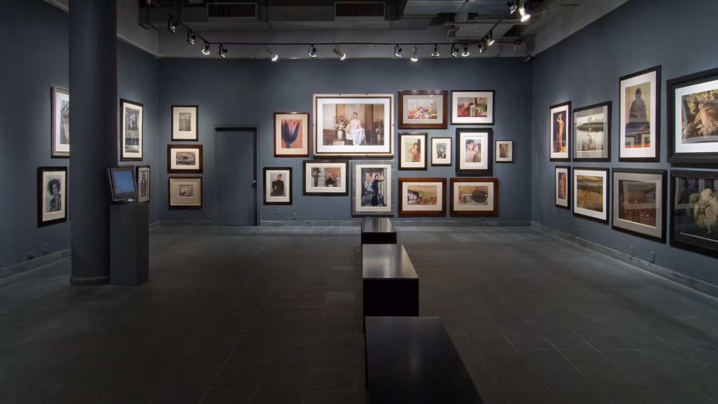 Wide-angle view of back gallery space at the Visual Arts Museum, showing framed photographs hung salon-style across three walls, with three black minimalist benches in the center of space.