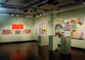 Photograph of illustrations on paper hung on green gallery walls, with three pedestals containing artist books in front of the wall to the left.