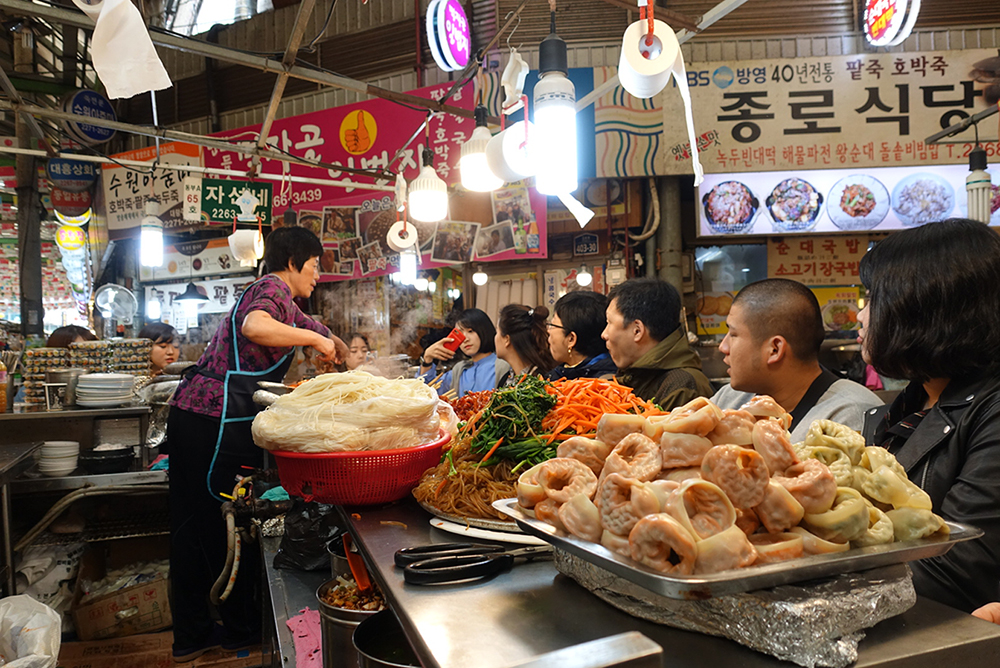 An image of seven people watching a woman cook behind a counter in South Korea.
