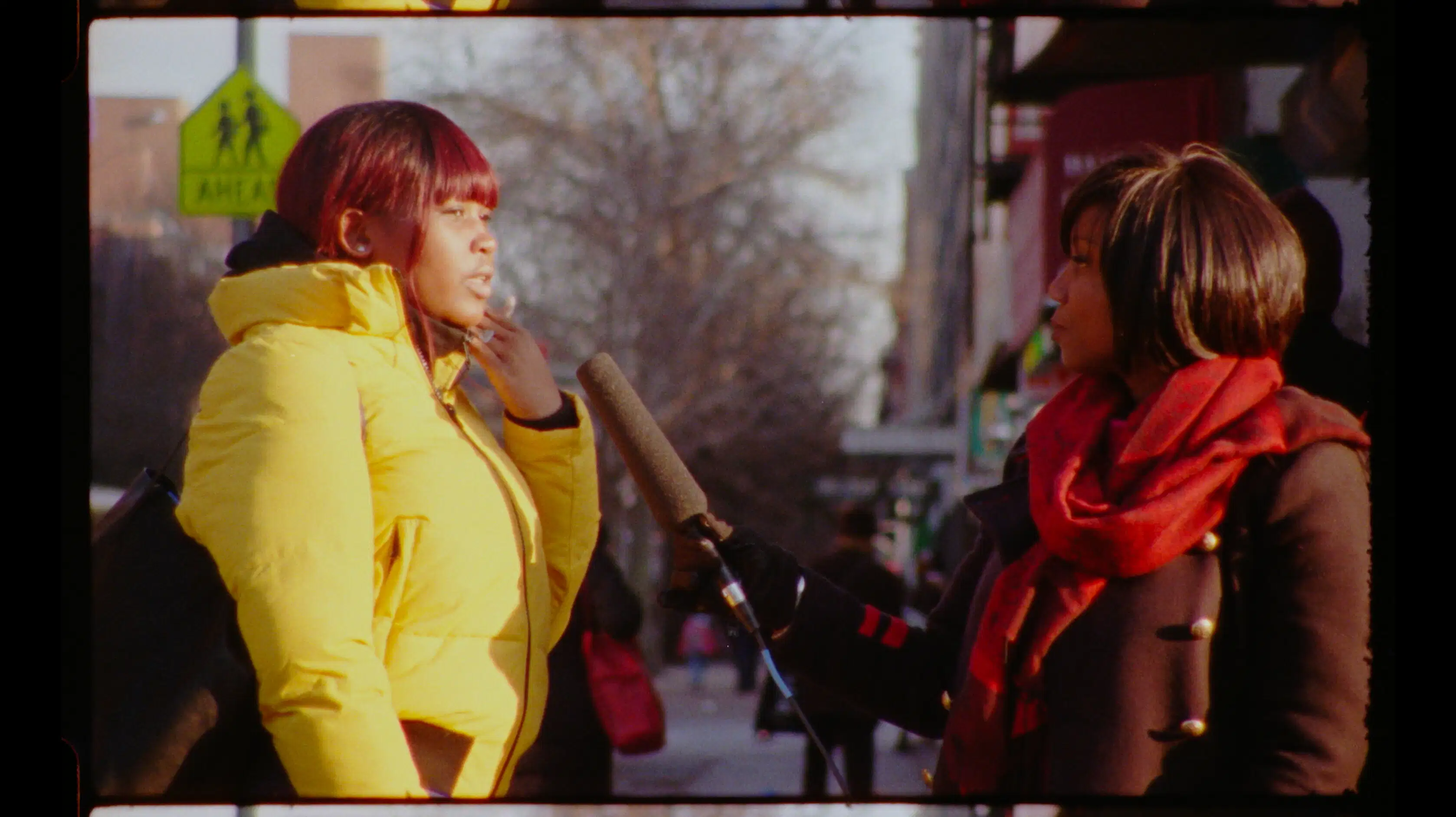 Video still of a woman interviewing another woman on the street. The interviewer hold a large microphone up. 