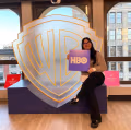 A student sits in front of a large illuminated Warner Bros. shield logo, holding a purple HBO sign and smiling. Two small signs on either side read “Warner Bros. Discovery Intern.