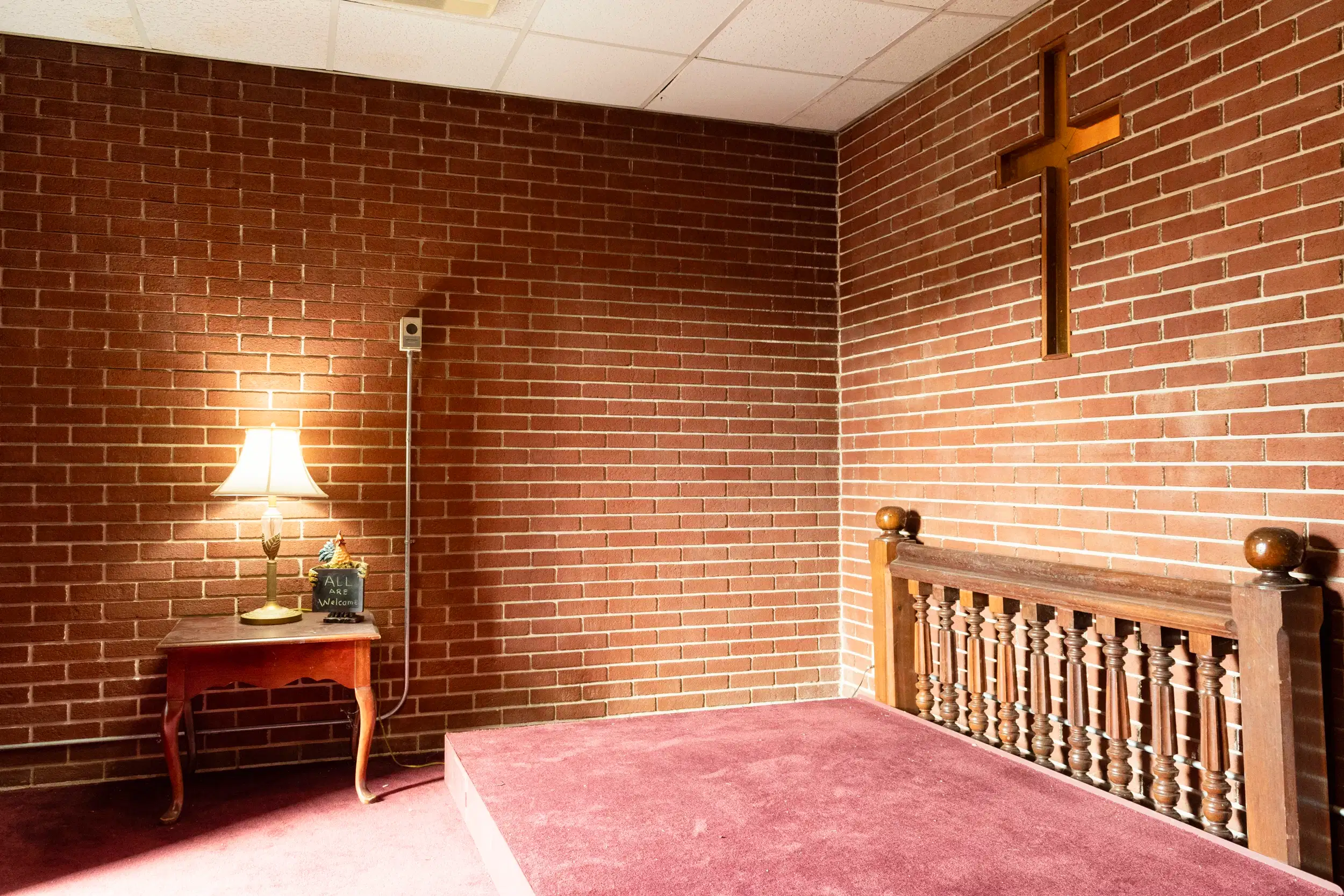 A small prayer room with an altar, side table, and lamp. A cross is embedded in the brick wall.