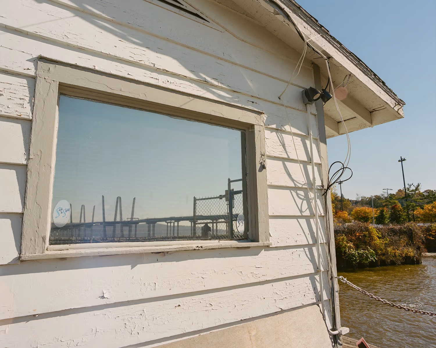 The window of a white shed frames a bridge stretching across a river with autumnal foliage in the background.