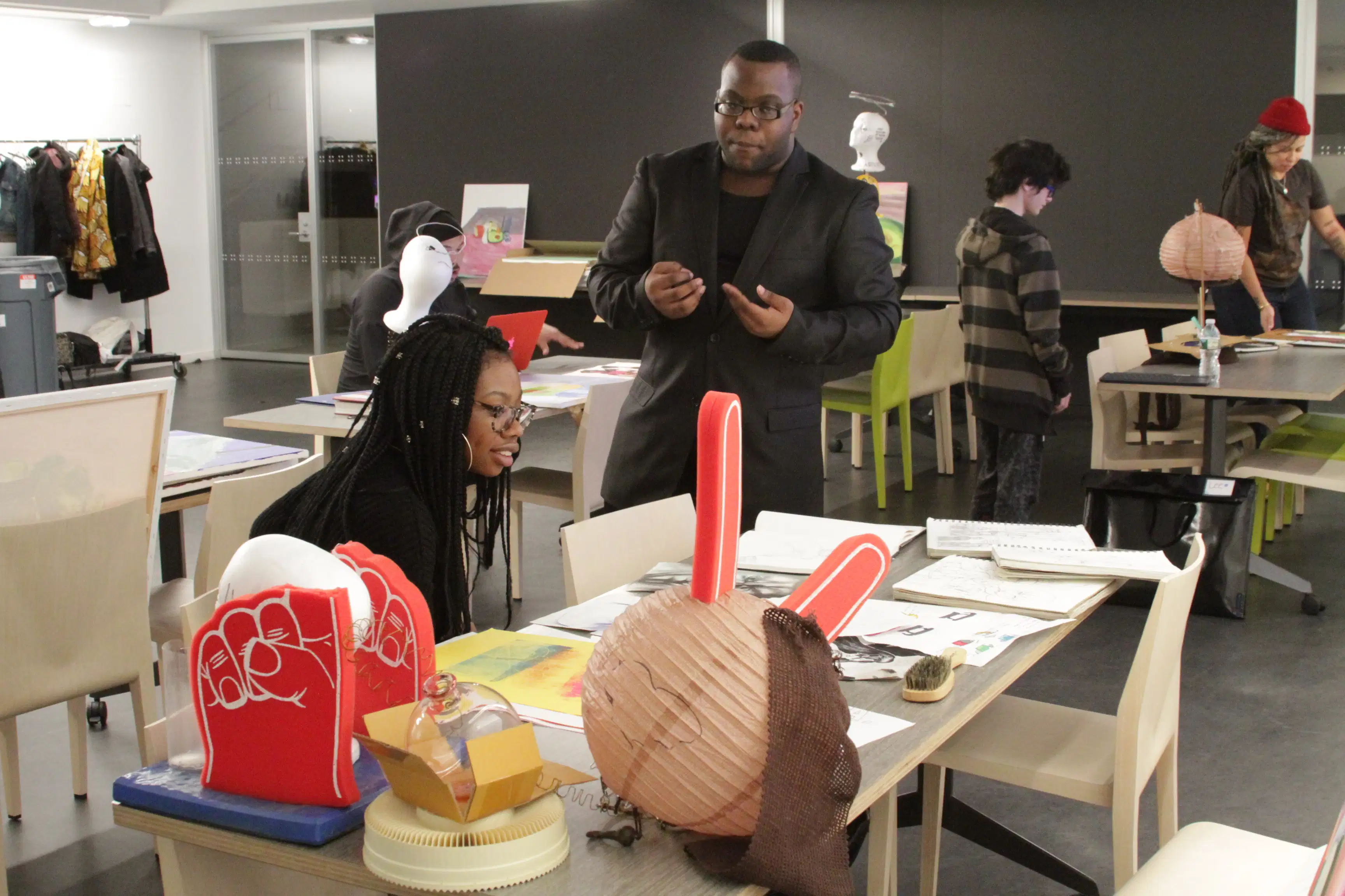 <p "="">A color photograph of several tables with two- and three-dimensional artworks on them. In the foreground, one person sits and looks at the art while another person is standing and gesturing.
