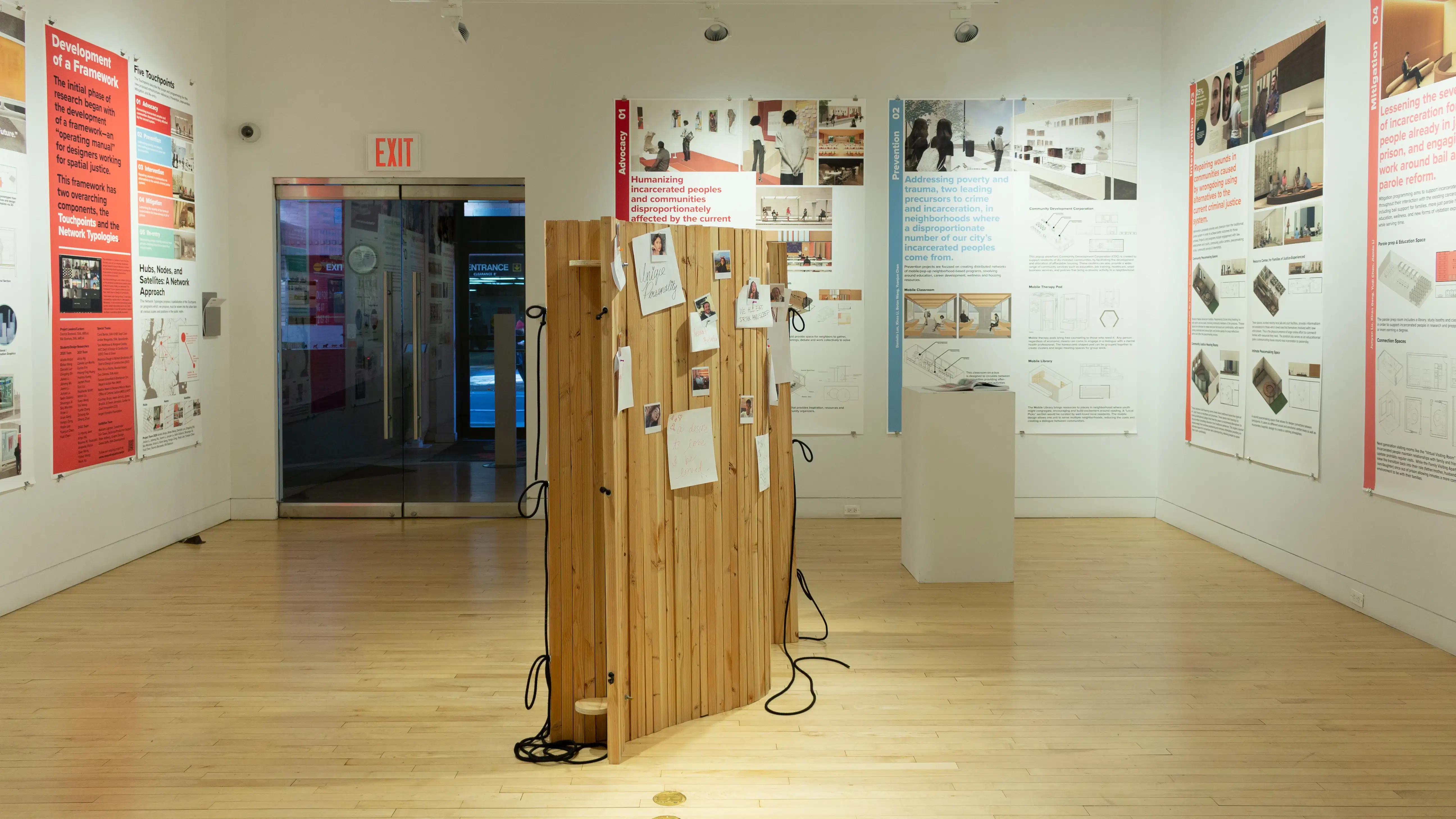 Exhibition view of An Infrastructure for Restorative Justice, featuring wooden sculpture, pedestal, and large posters on the walls.