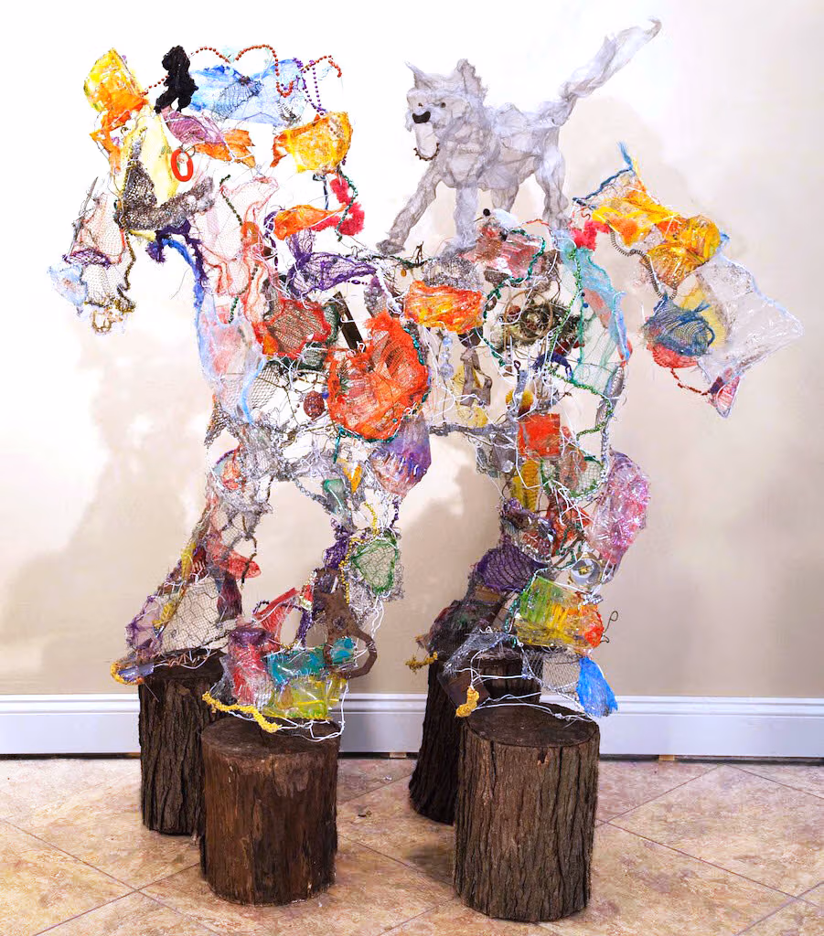 A colorful wire sculpture in the shape of a horse with a cat on top of it standing on four tree stumps.