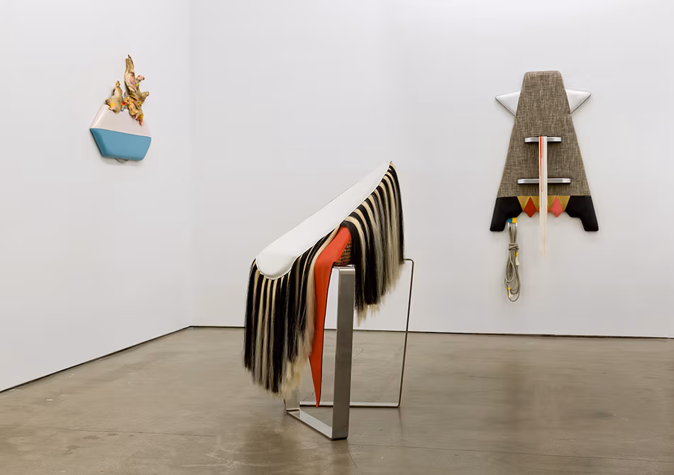 <p "="">An installation view of three fabric-based sculptures—two hung on adjacent gallery walls and one tilting on a metal stand on the concrete floor.
