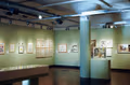 Photograph of gallery space with green walls and many framed illustrations, along with a large vitrine in the center of the room.