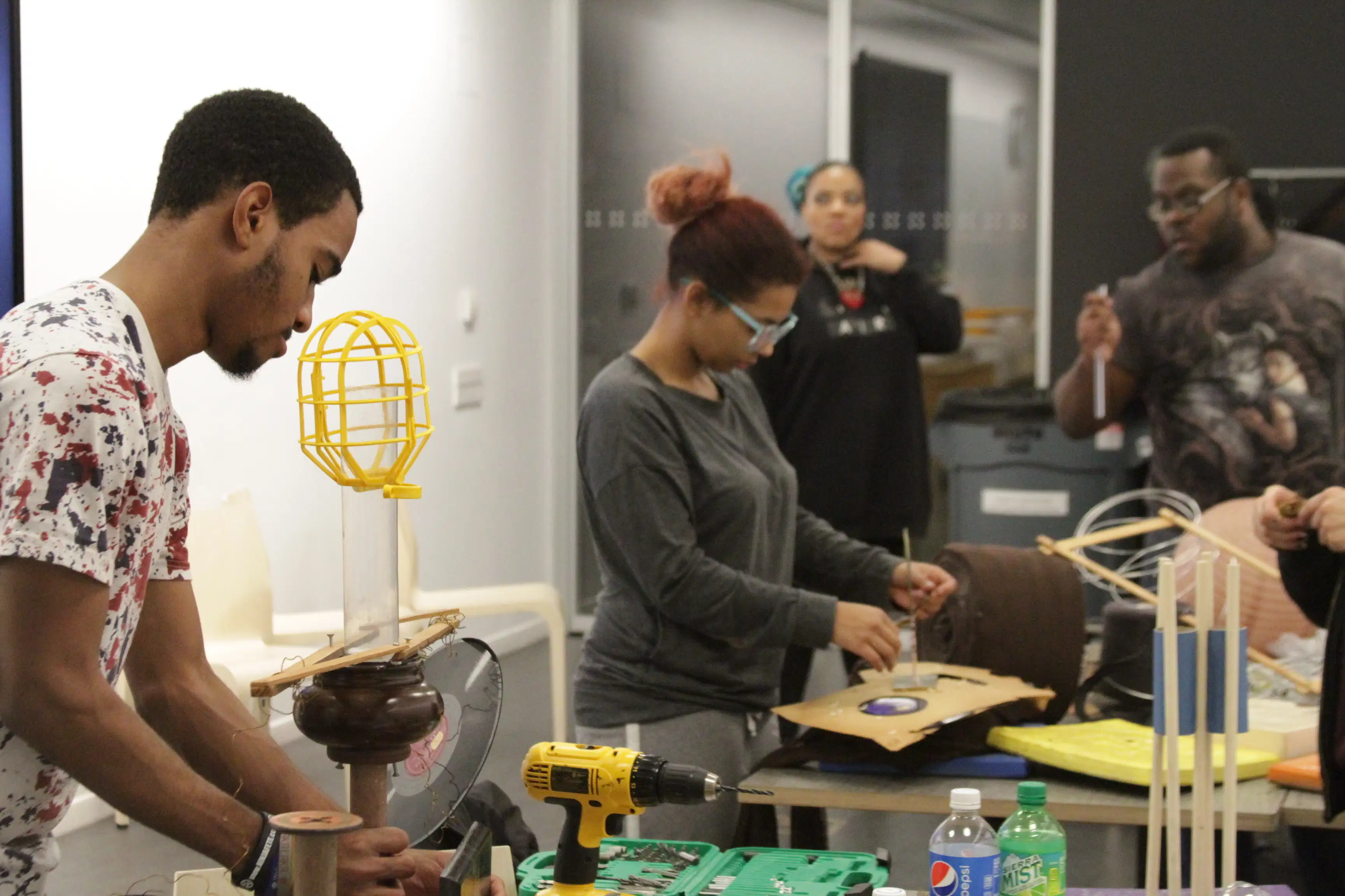 <p "="">Four students stand at or near tables with a variety of art-making materials and tools on them.
