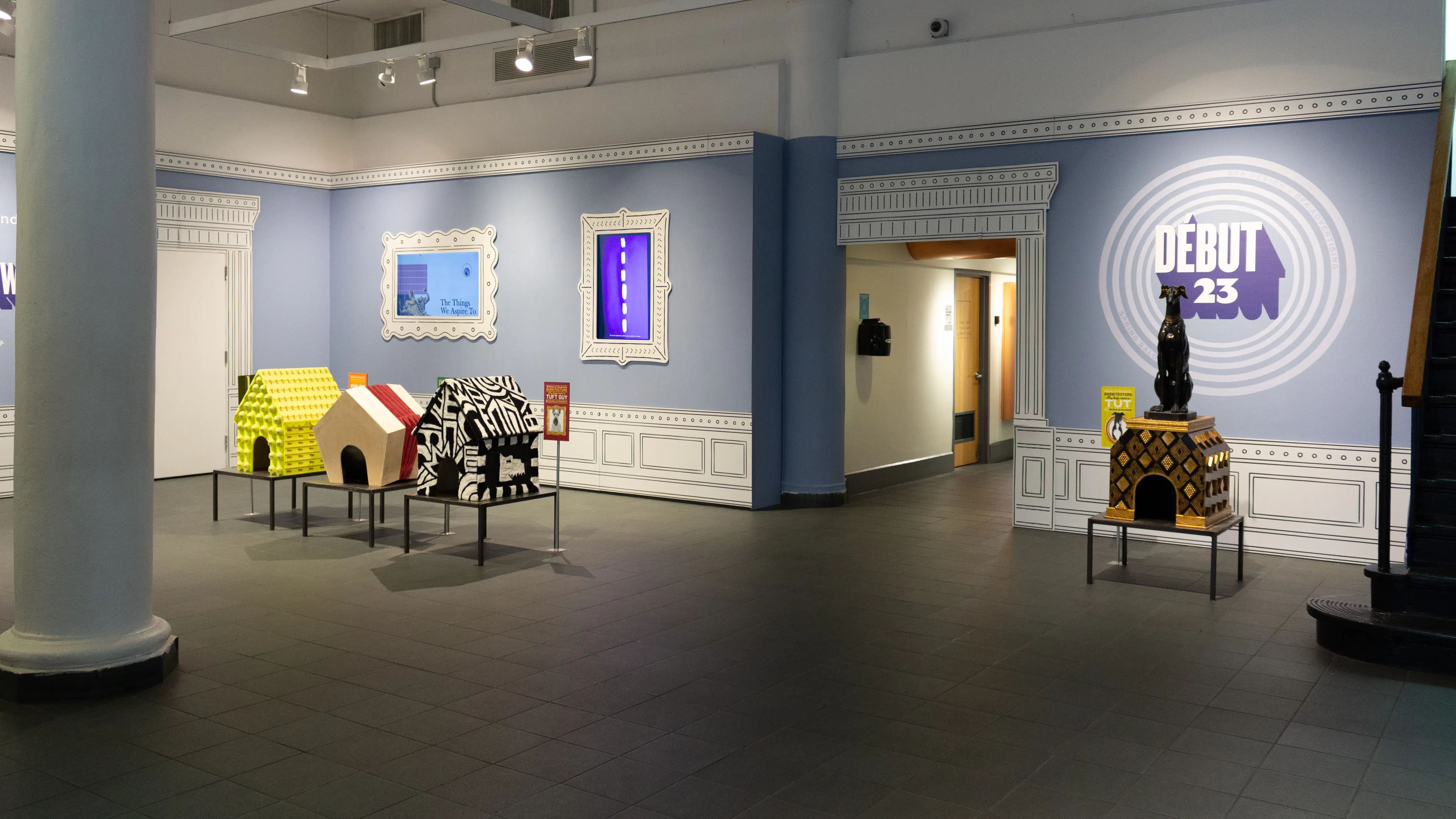 Installation image featuring a large room with dog house sculptures in the center and framed monitors on the walls.