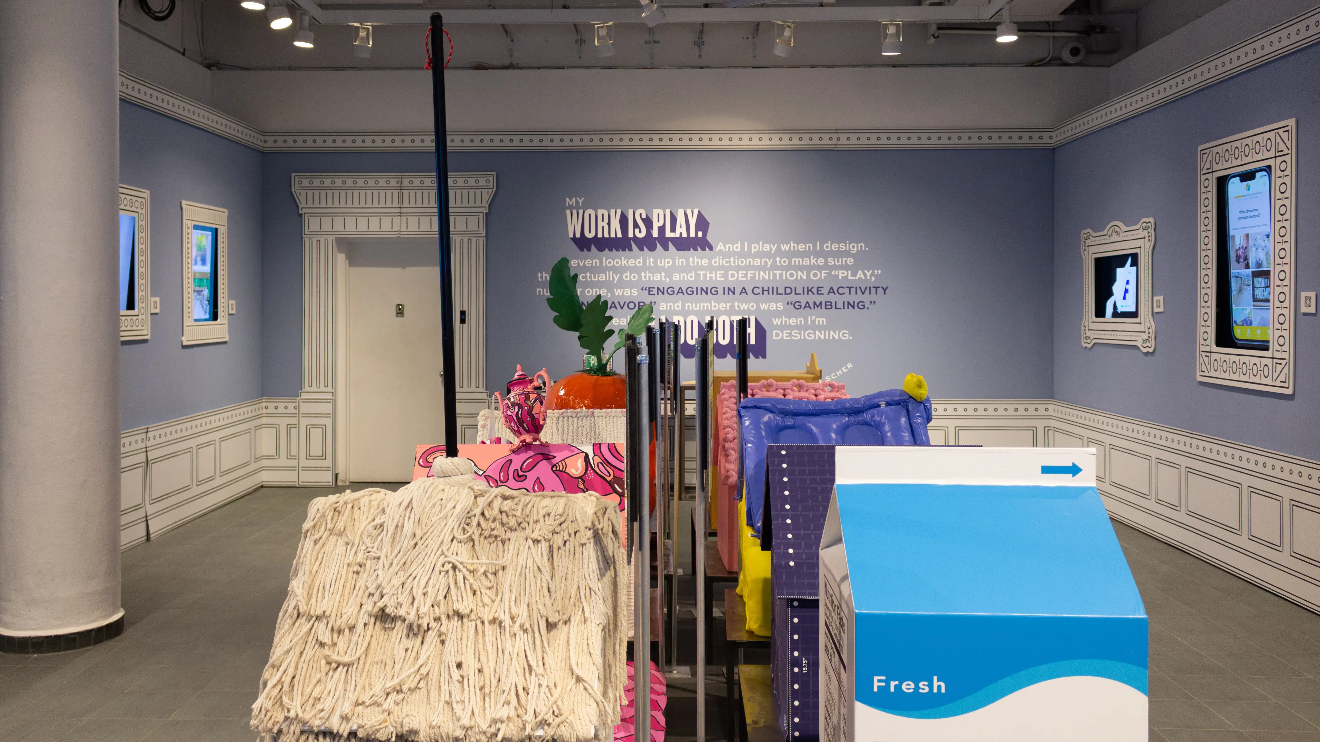 Installation image featuring a large room with ten dog house sculptures in the center and four visible framed monitors on the walls.