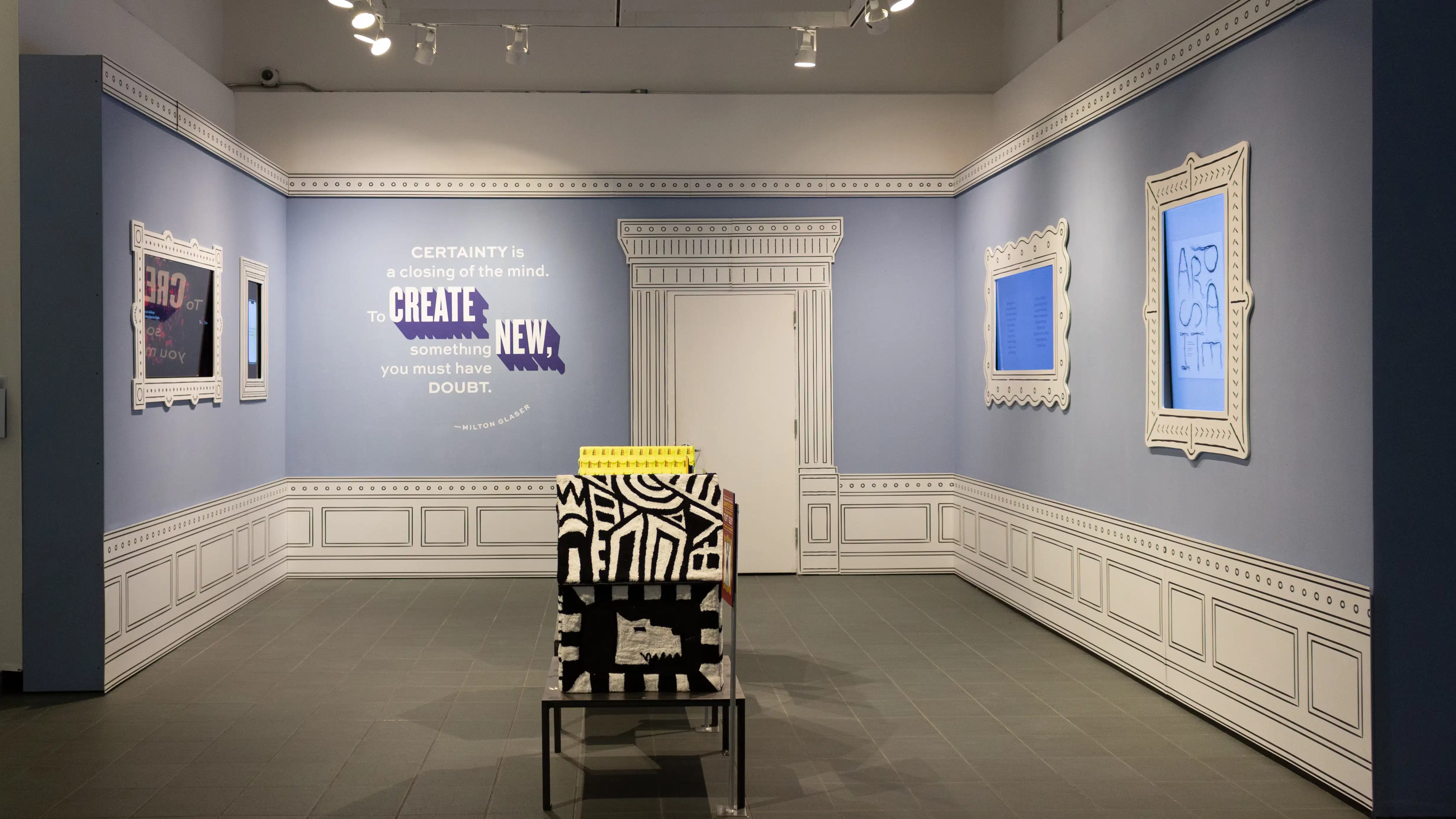 Installation image featuring a large room with three dog house sculptures in the center and four framed monitors on the walls.