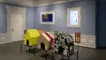 Installation image featuring a large room with three dog house sculptures in the center and two framed monitors on the walls.