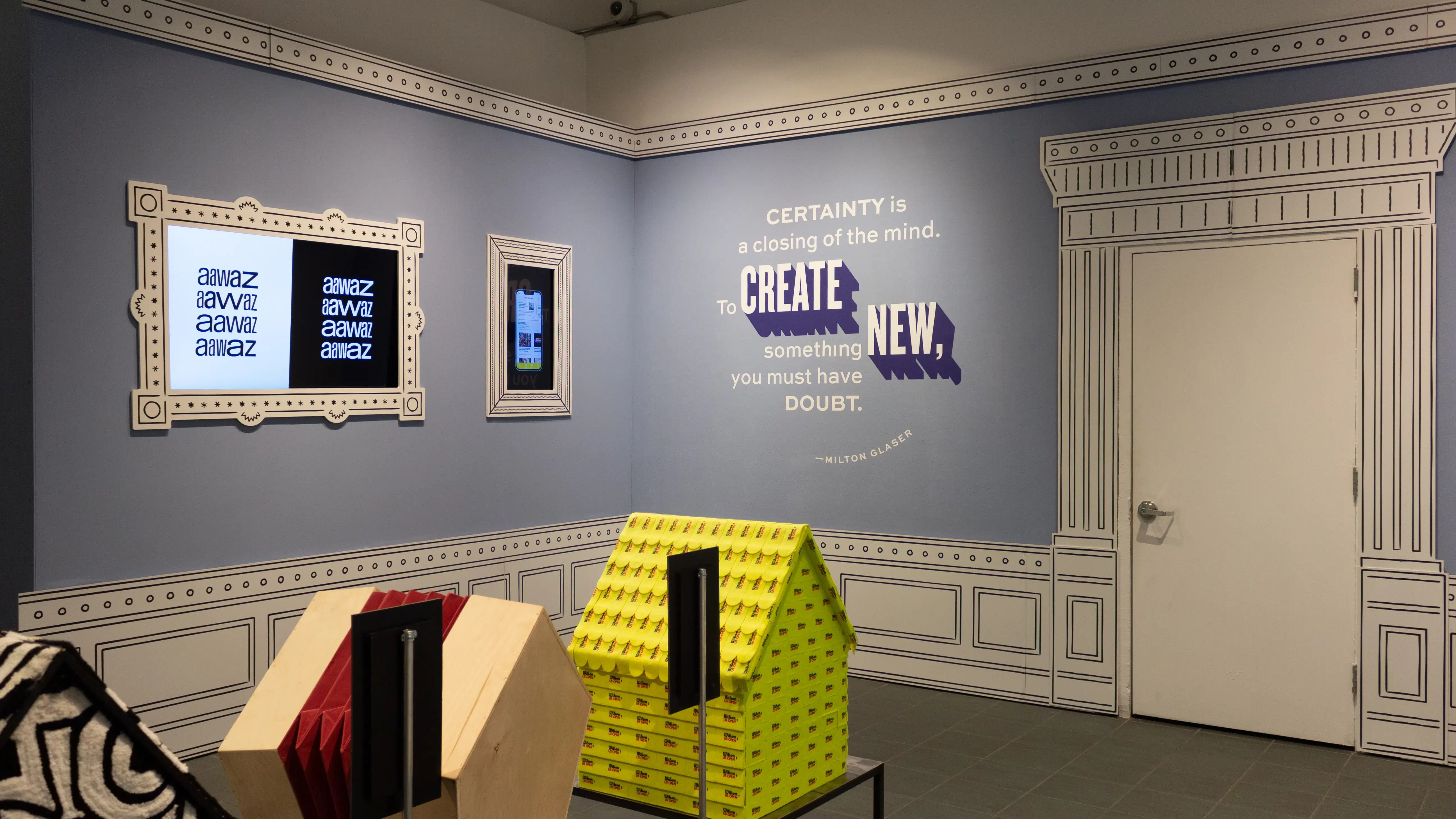 Installation image featuring a large room with three dog house sculptures in the center and two framed monitors on the walls.