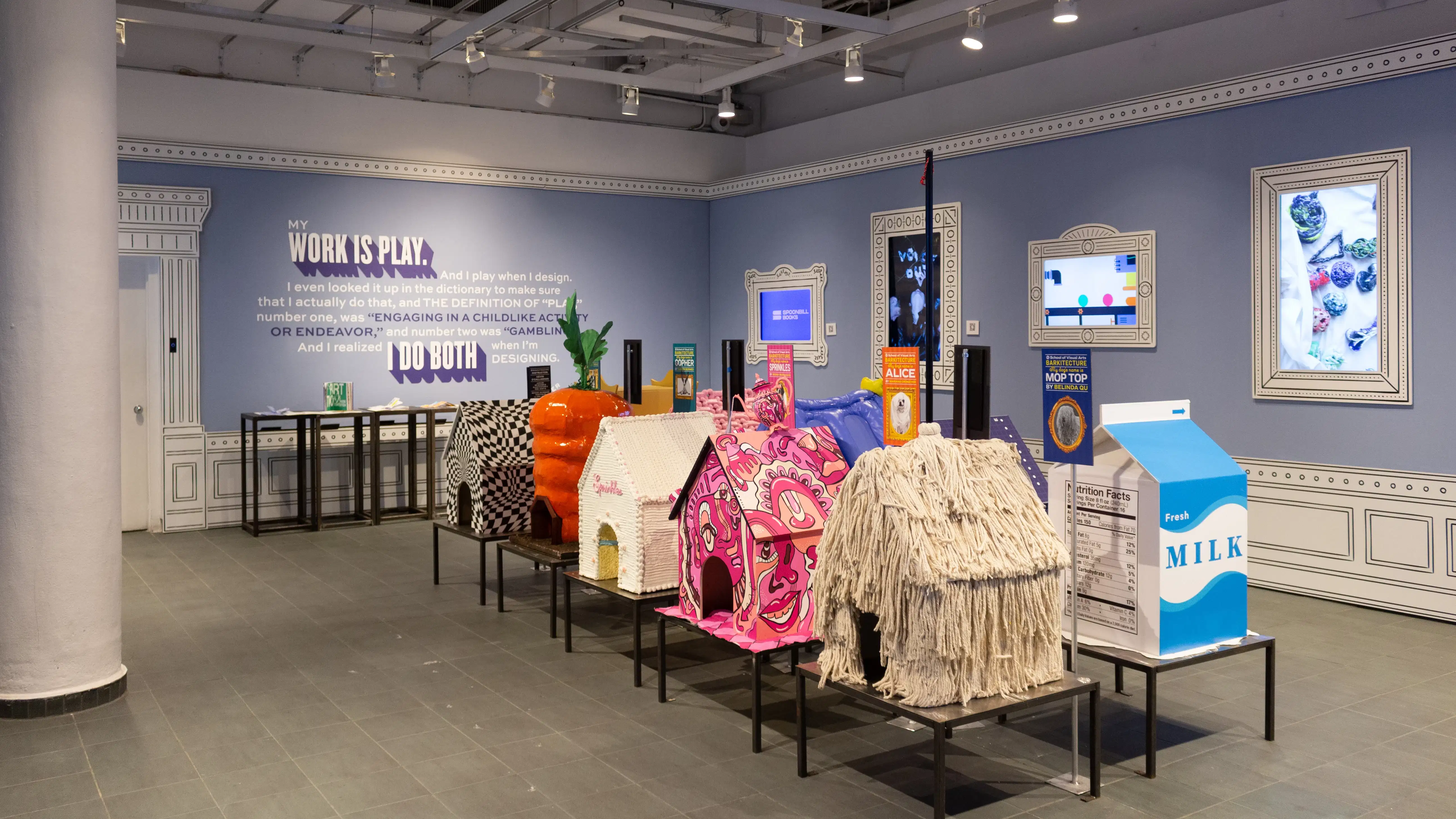 Installation image featuring a large room with ten dog house sculptures in the center and four visible framed monitors on the walls.