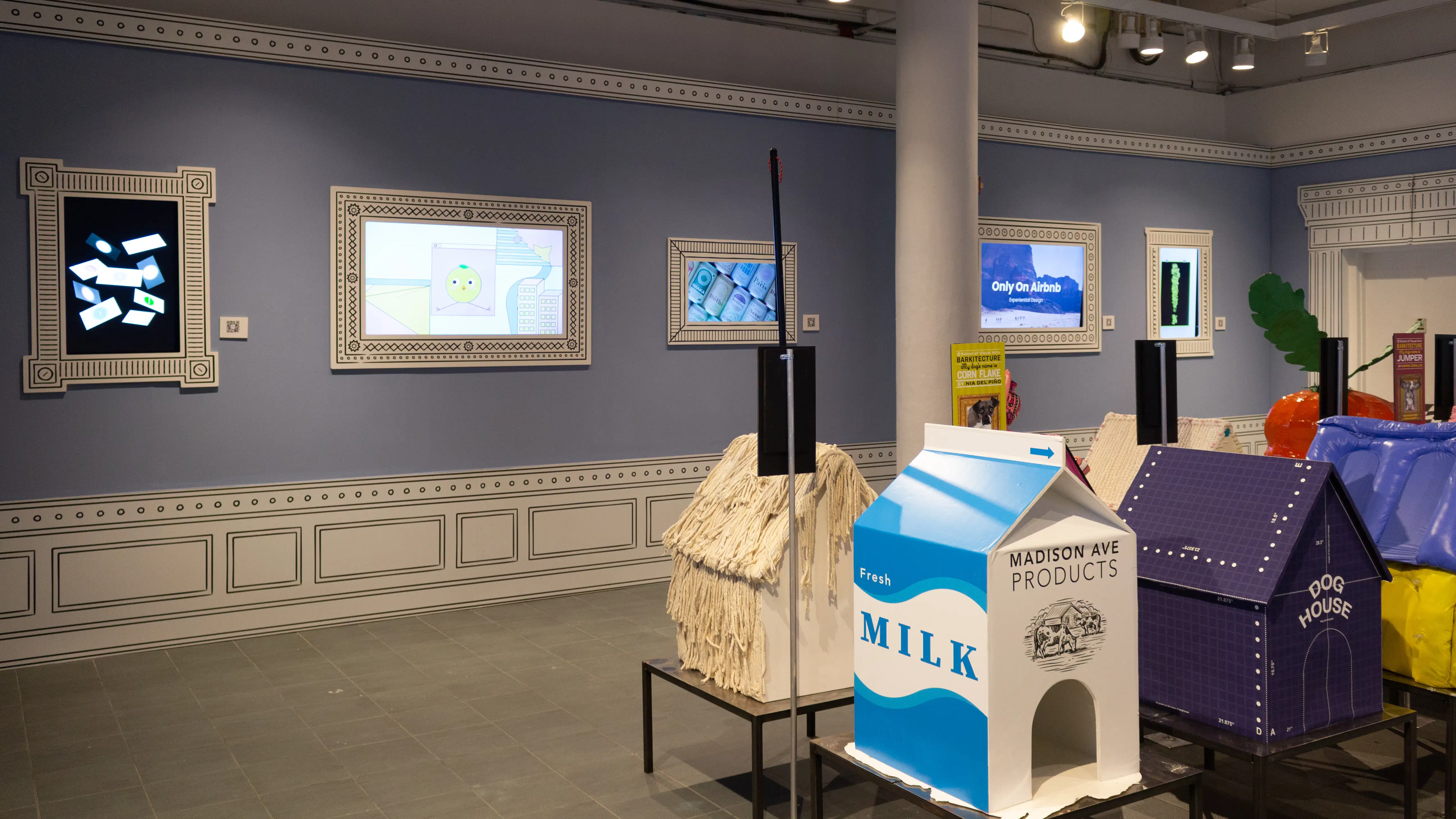 Installation image featuring a large room with seven dog house sculptures in the center and five framed monitors on the walls.