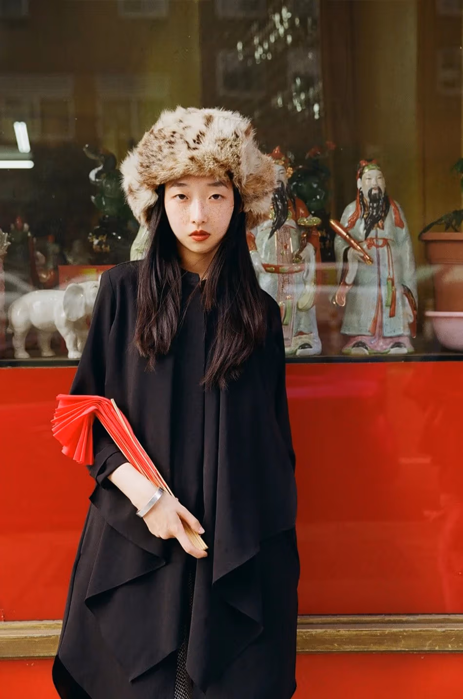 Portrait of a young woman with a big fuzzy hat posing in front of a glass window displaying porcelain sculptures. 