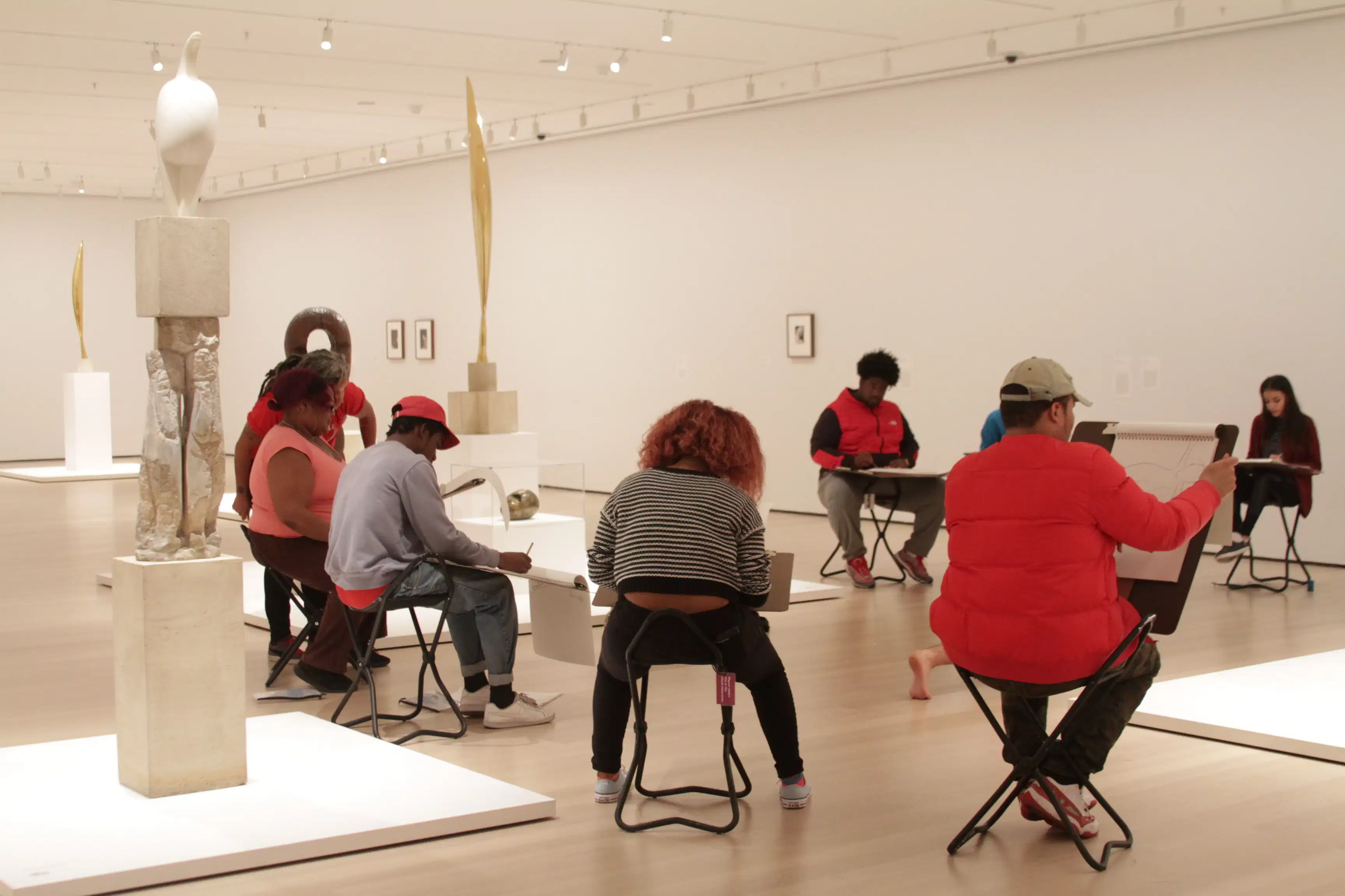 <p "="">A photograph of several art students sitting and sketching amid the sculptures and art on view in a white-walled gallery space.
