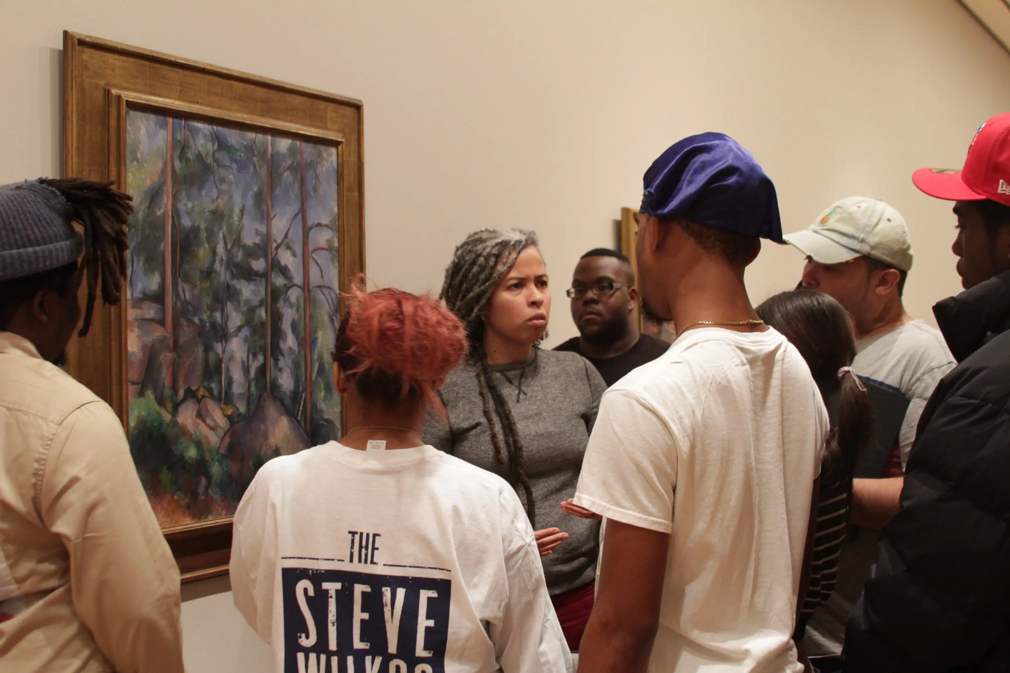 A woman stands next to a painting of a forest scene and talks to a group of people standing around her.
