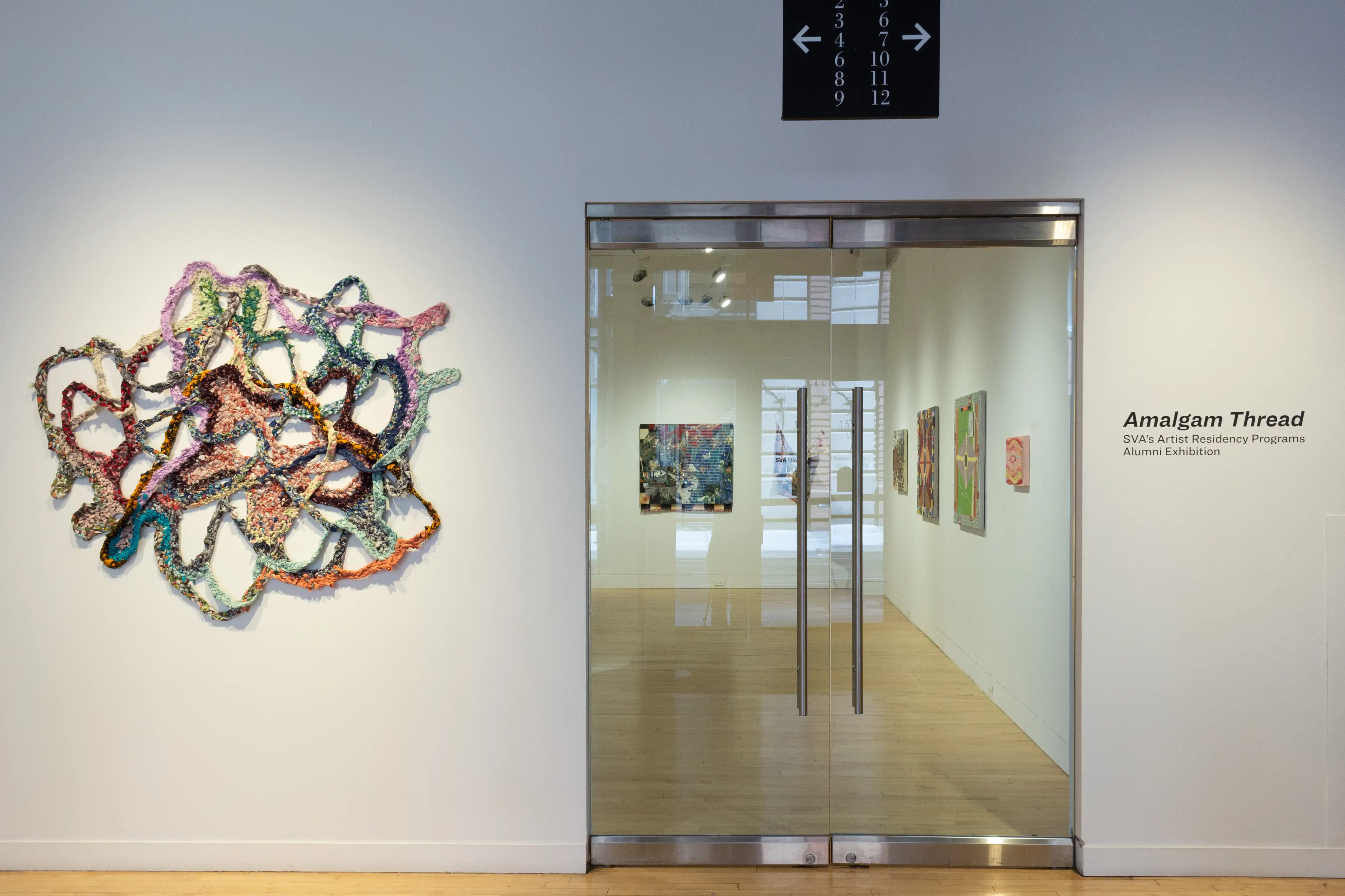 Installation image featuring a textile work and the gallery doors, with wall vinyl reading, "Amalgam Thread" to the right.