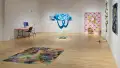 Art gallery filled with work across three walls, including a carpet on the floor and a suspended rain cloud sculpture. 