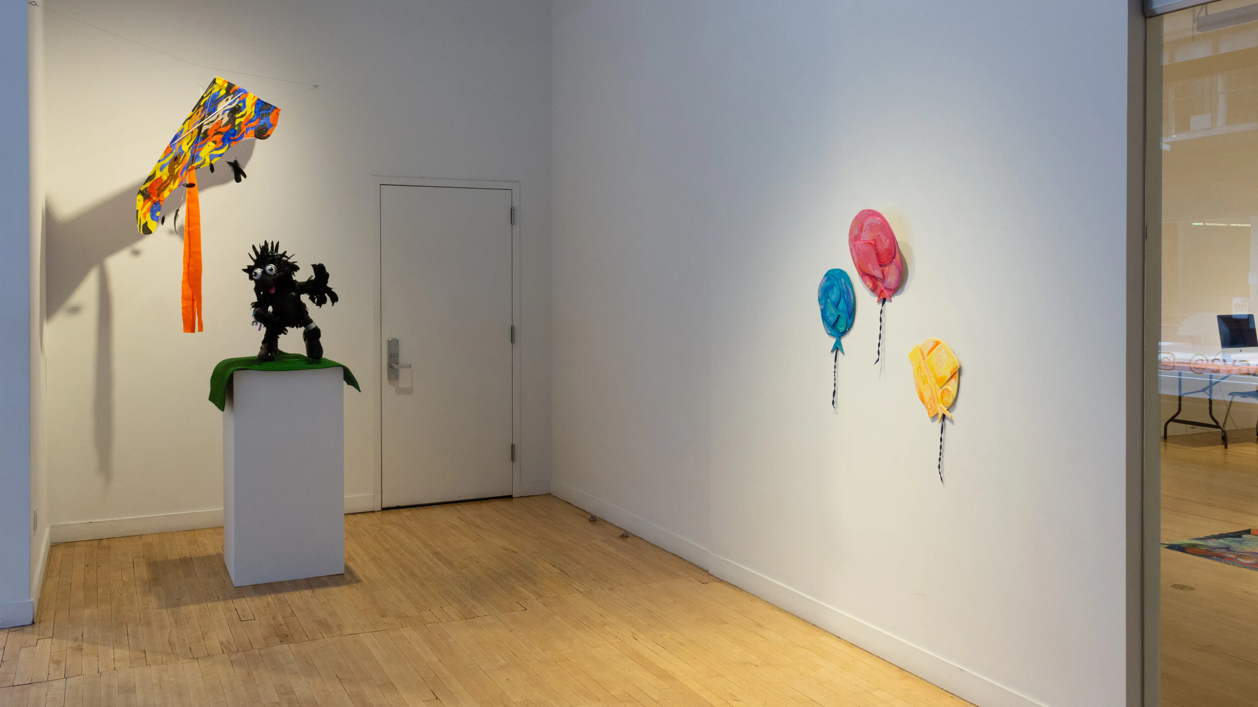 On the far left a kite hangs suspended between two walls, underneath it is a sculpture of a human figure on top of a pedestal, and to the far right are three balloon sculptures. 