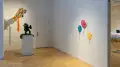 On the far left a kite hangs suspended between two walls, underneath it is a sculpture of a human figure on top of a pedestal, and to the far right are three balloon sculptures. 