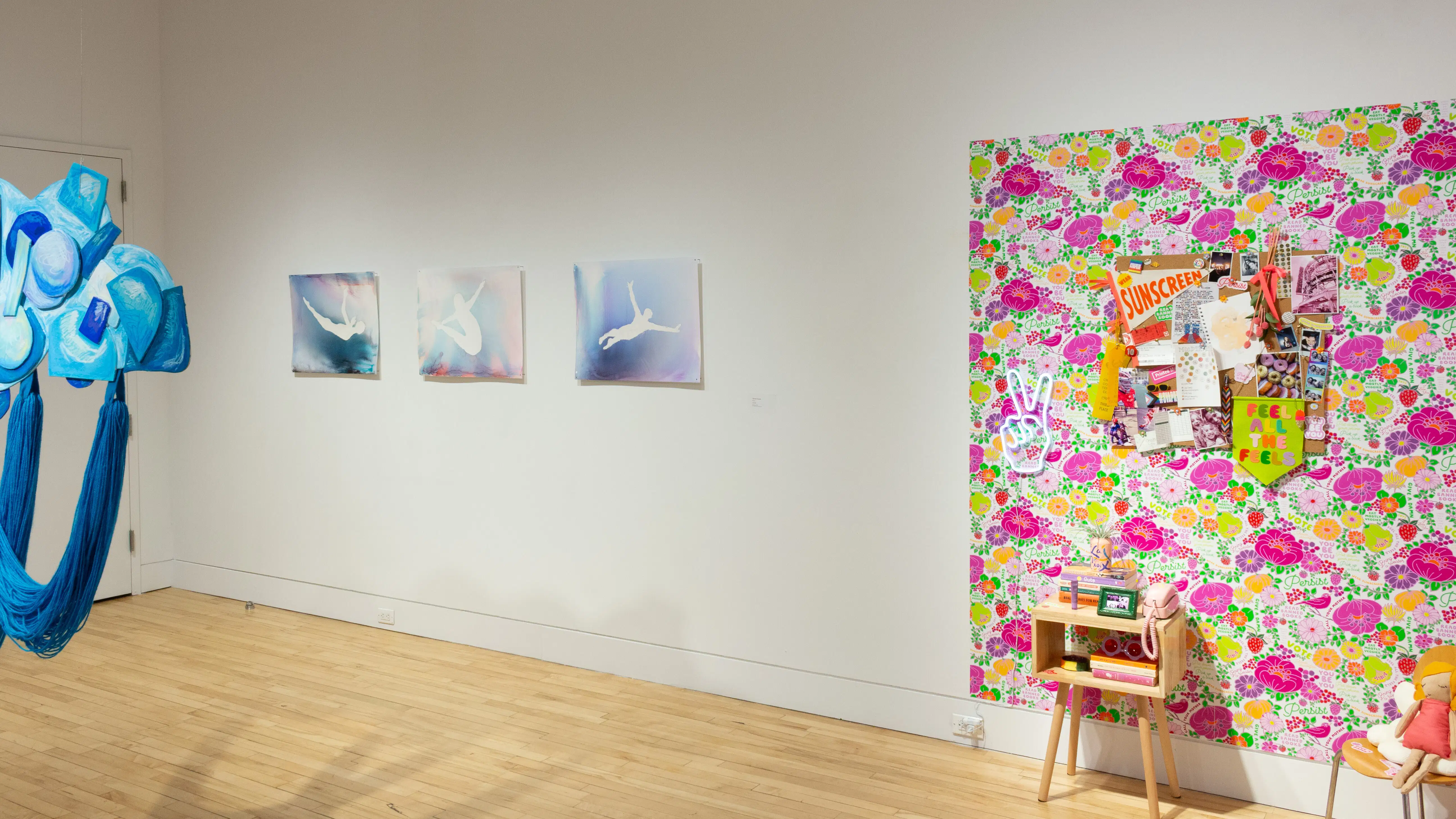 On the left, three artworks hang from a wall depict a person in mid-air, and on the far right, an artwork installation depicts an interior bedroom-like space with floral wallpaper.