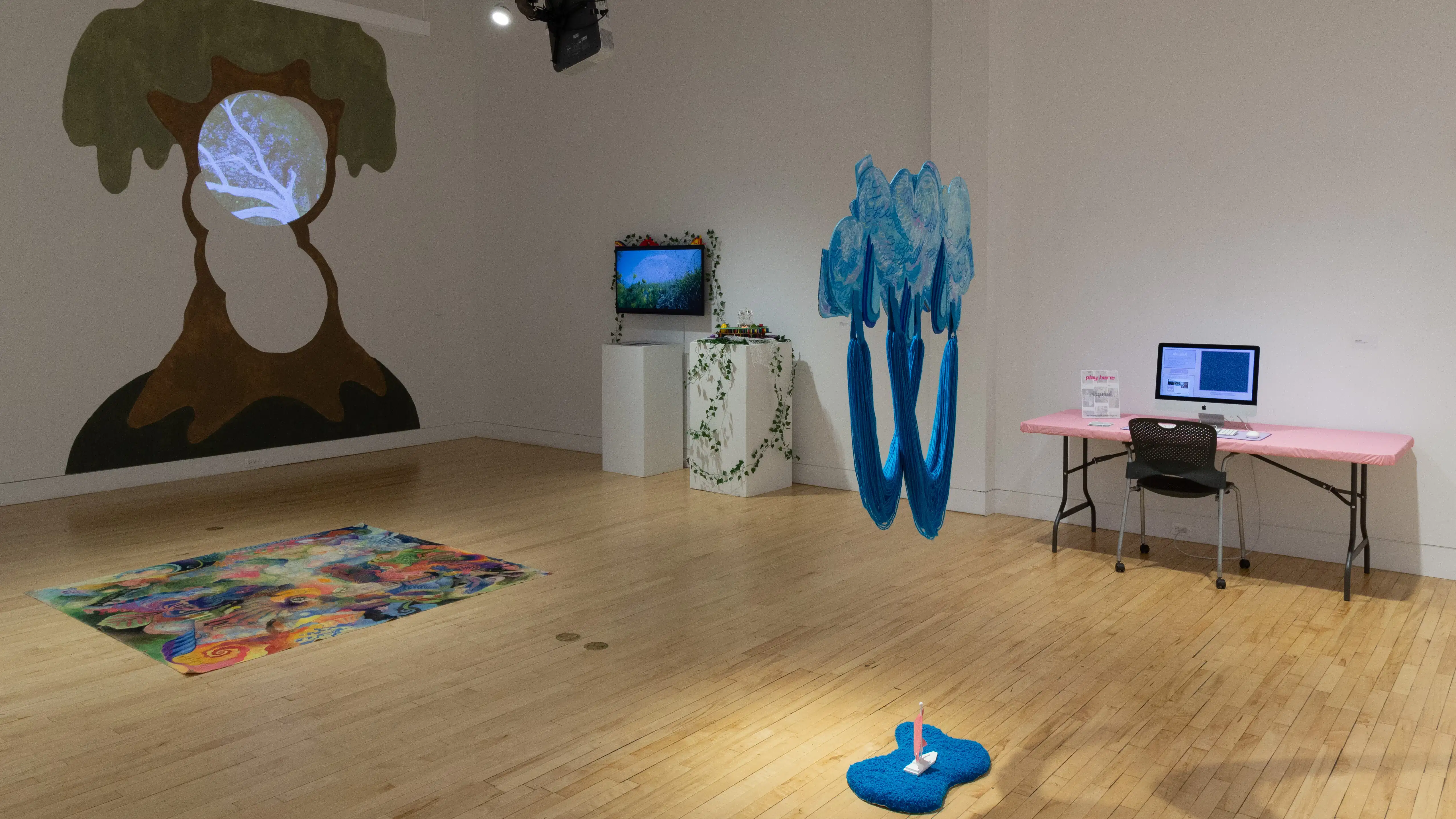  On the left wall is a large tree painting, in front of it is a colorful rug, and on the far right are two screens displaying artwork.