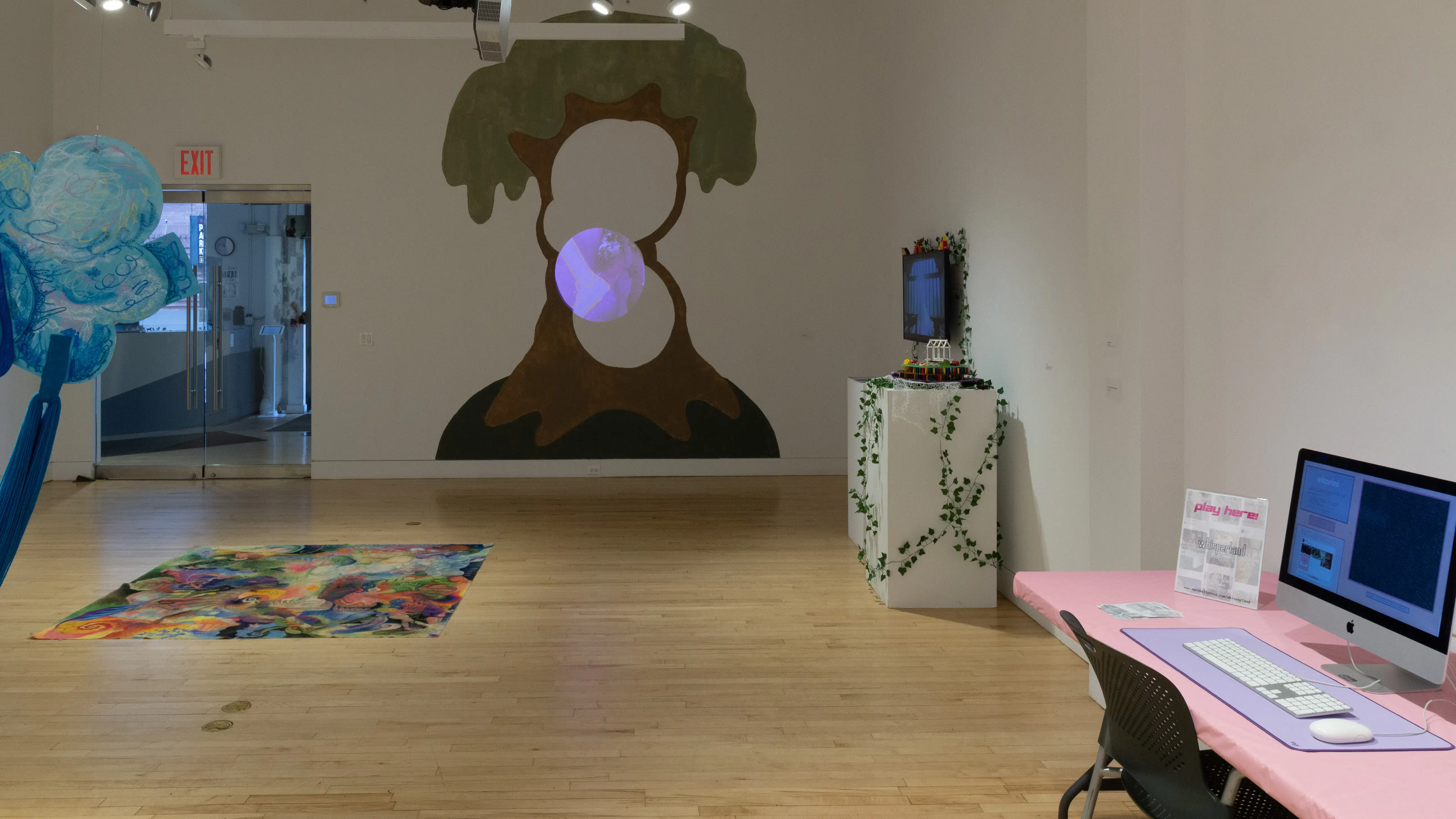 On the back wall is a large tree painting, in front of it is a colorful rug, and on the far right are two screens displaying artwork.