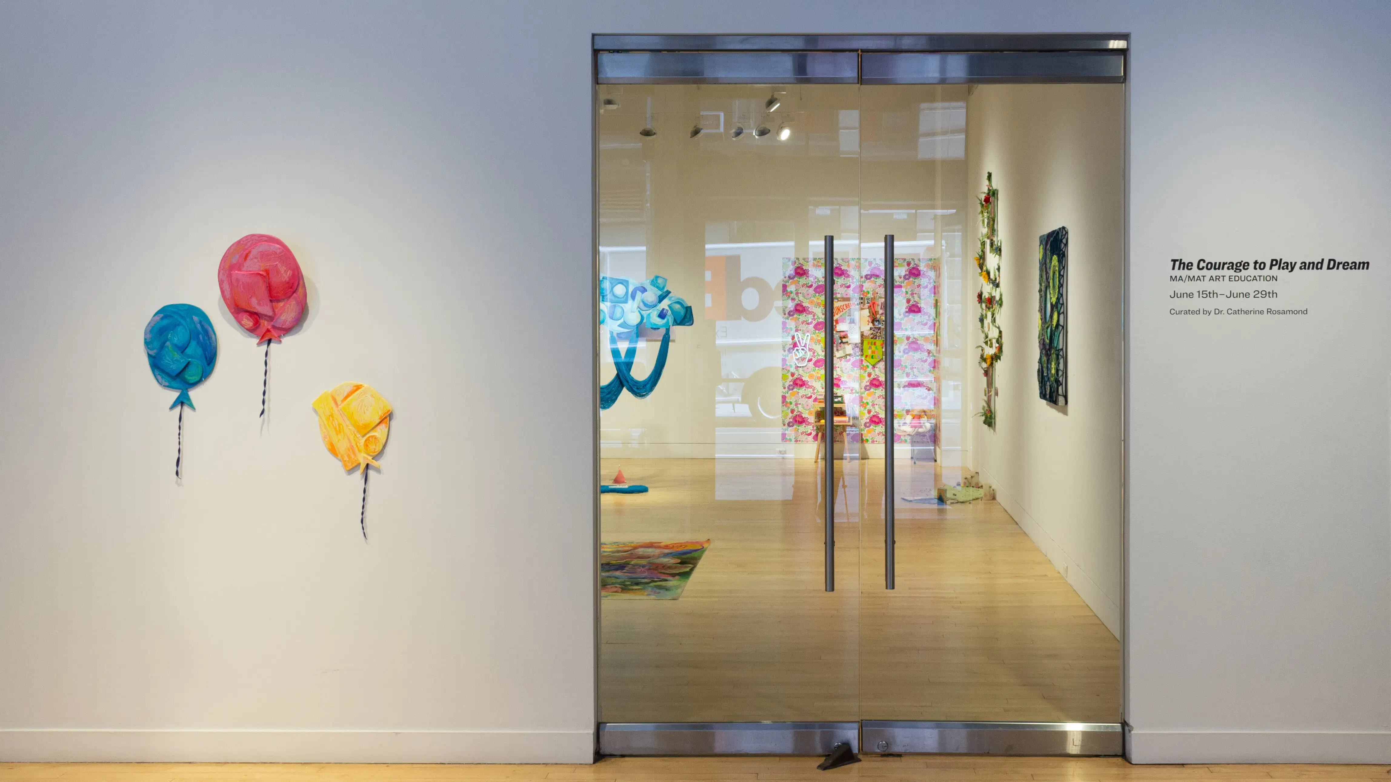 On the far left are three balloon sculptures attached to the wall, in the center is a view into an art gallery behind glass doors, and on the far left is the exhibition title on the wall.
