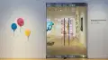 On the far left are three balloon sculptures attached to the wall, in the center is a view into an art gallery behind glass doors, and on the far left is the exhibition title on the wall.