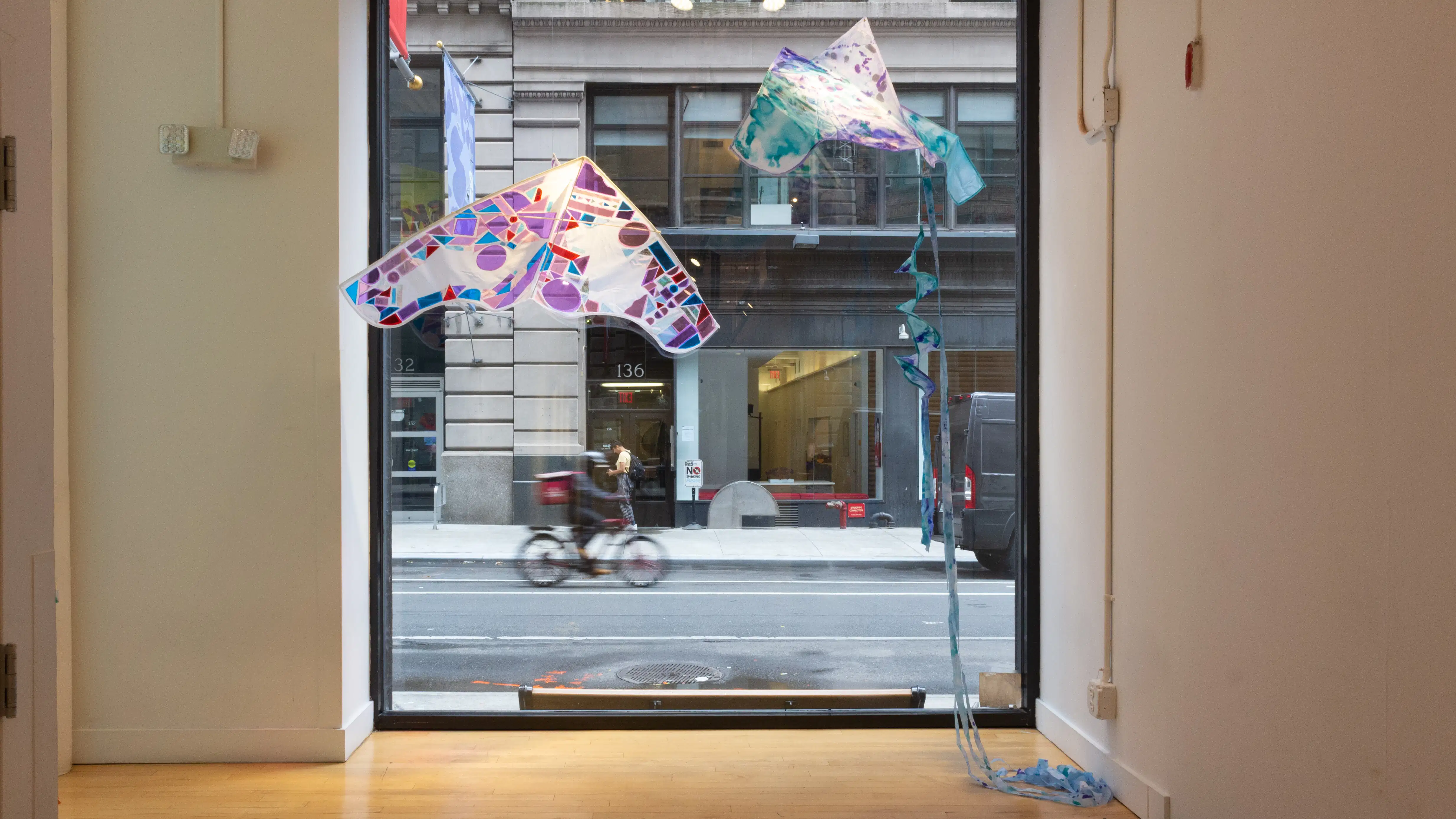 View into the street through a large window, in front of which two colorful kites are suspended.