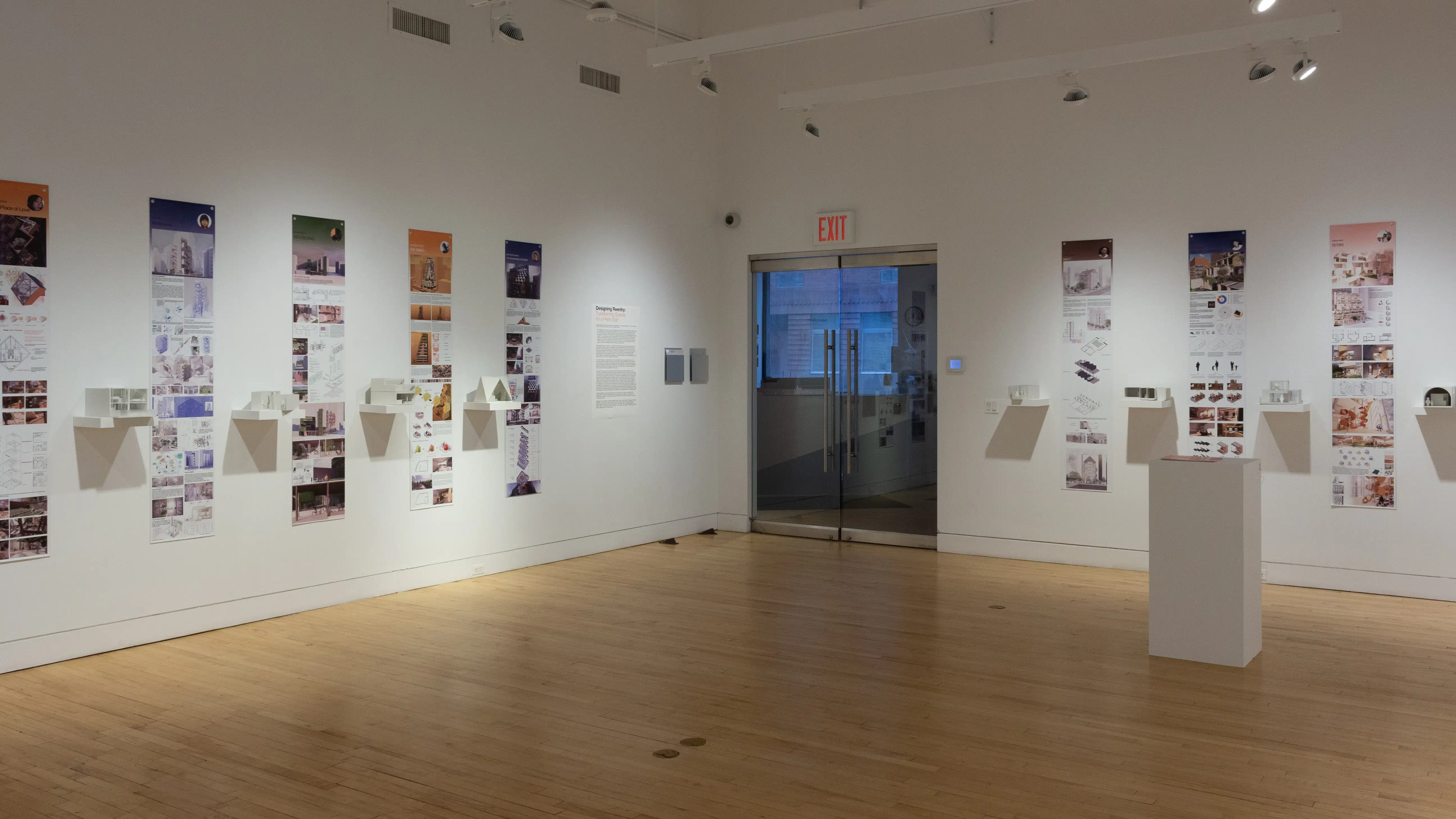 Installation image showing a three walls of a large gallery with building models on shelves, and information banners for each to the right. There is a pedestal on the floor of the gallery and glass doors on the back wall.