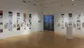 Installation image showing a three walls of a large gallery with building models on shelves, and information banners for each to the right. There is a pedestal on the floor of the gallery and glass doors on the back wall.