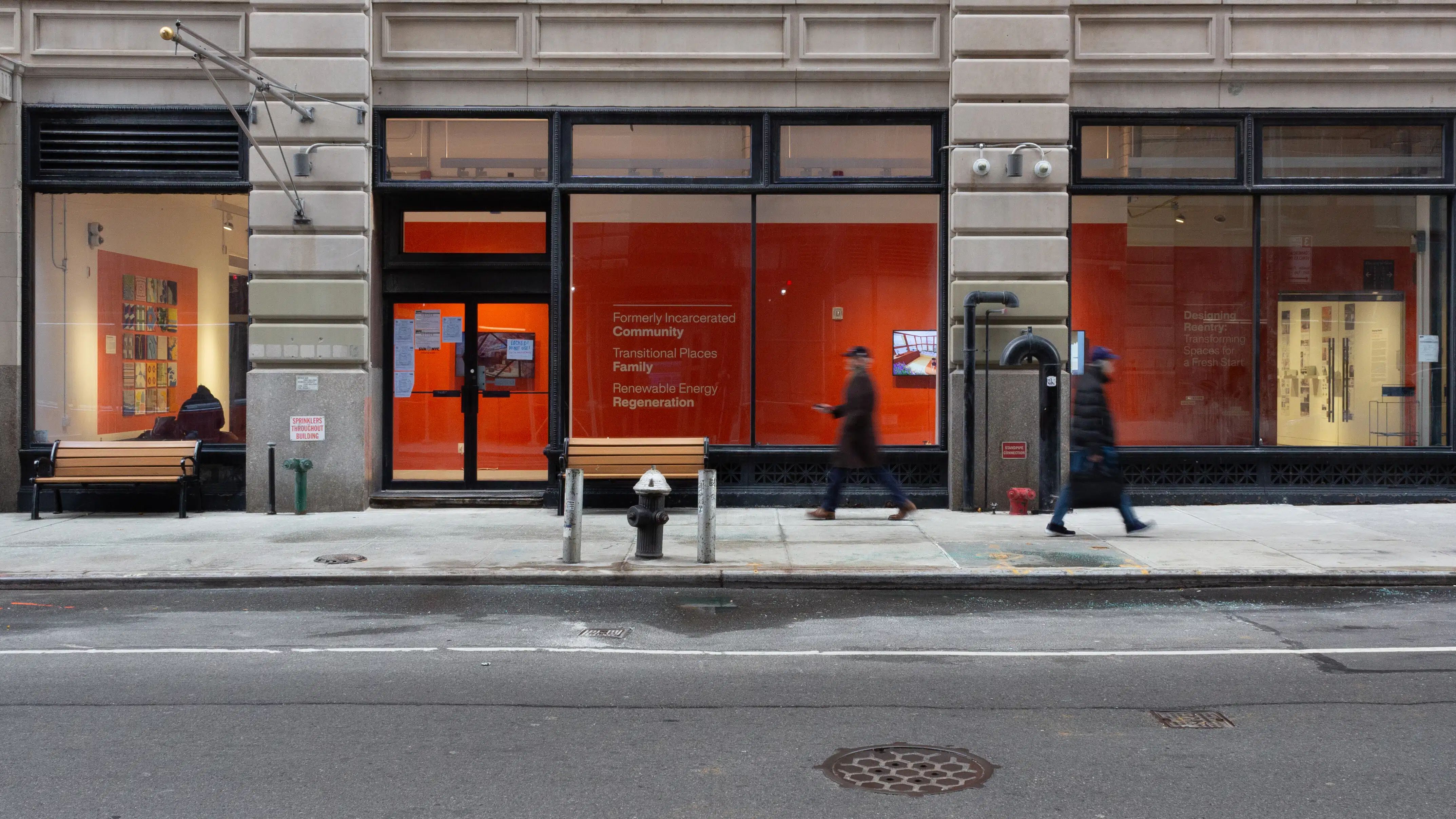 Street view of large windows showing a long gallery hallway with orange walls and monitors. Two people cross on the sidewalk in front.