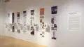 Installation image showing an angled view of a gallery wall with seven building models on shelves, and information banners for each to the right.