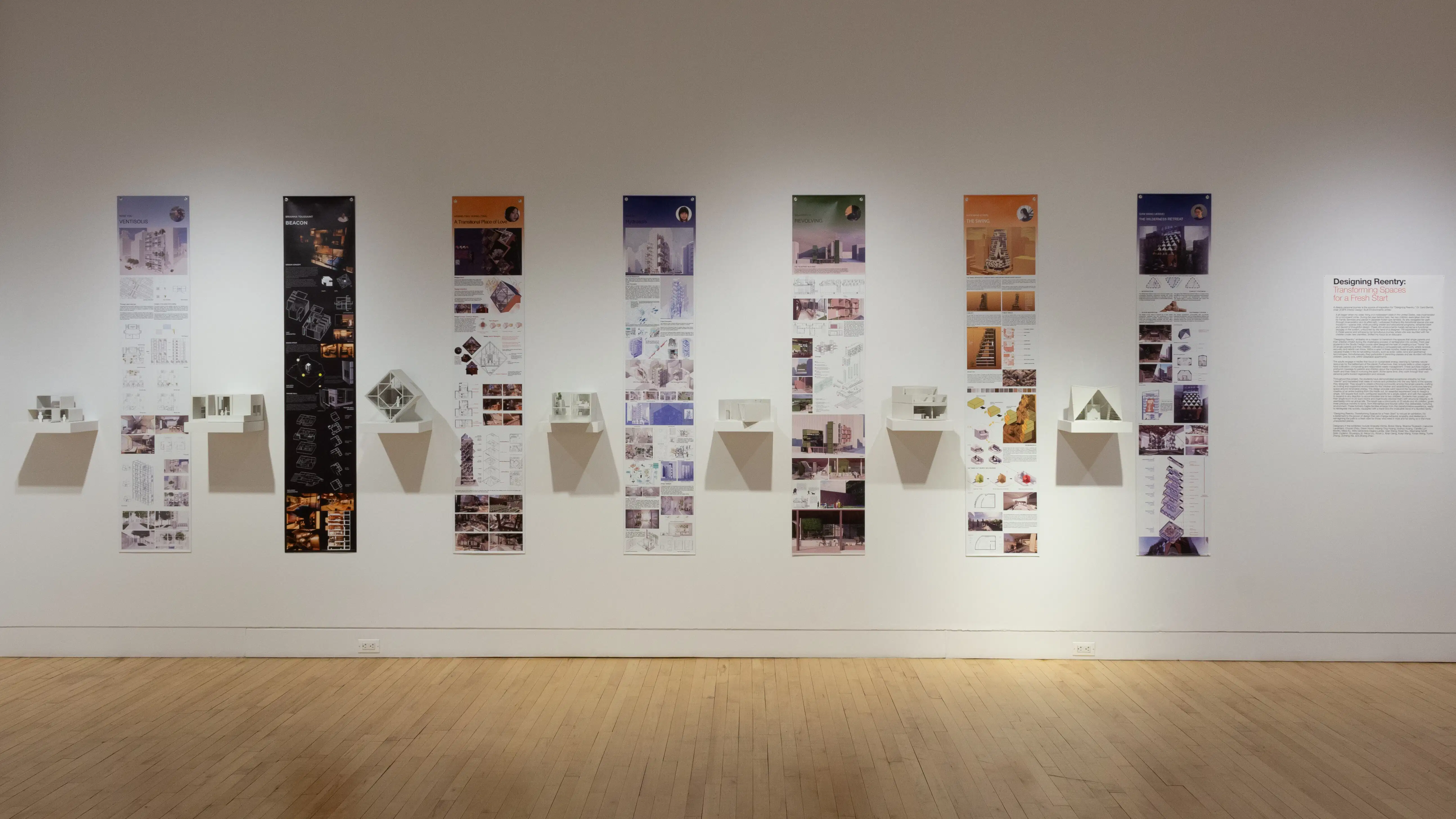 Installation image showing a straight view of a gallery wall with seven building models on shelves, and information banners for each to the right.