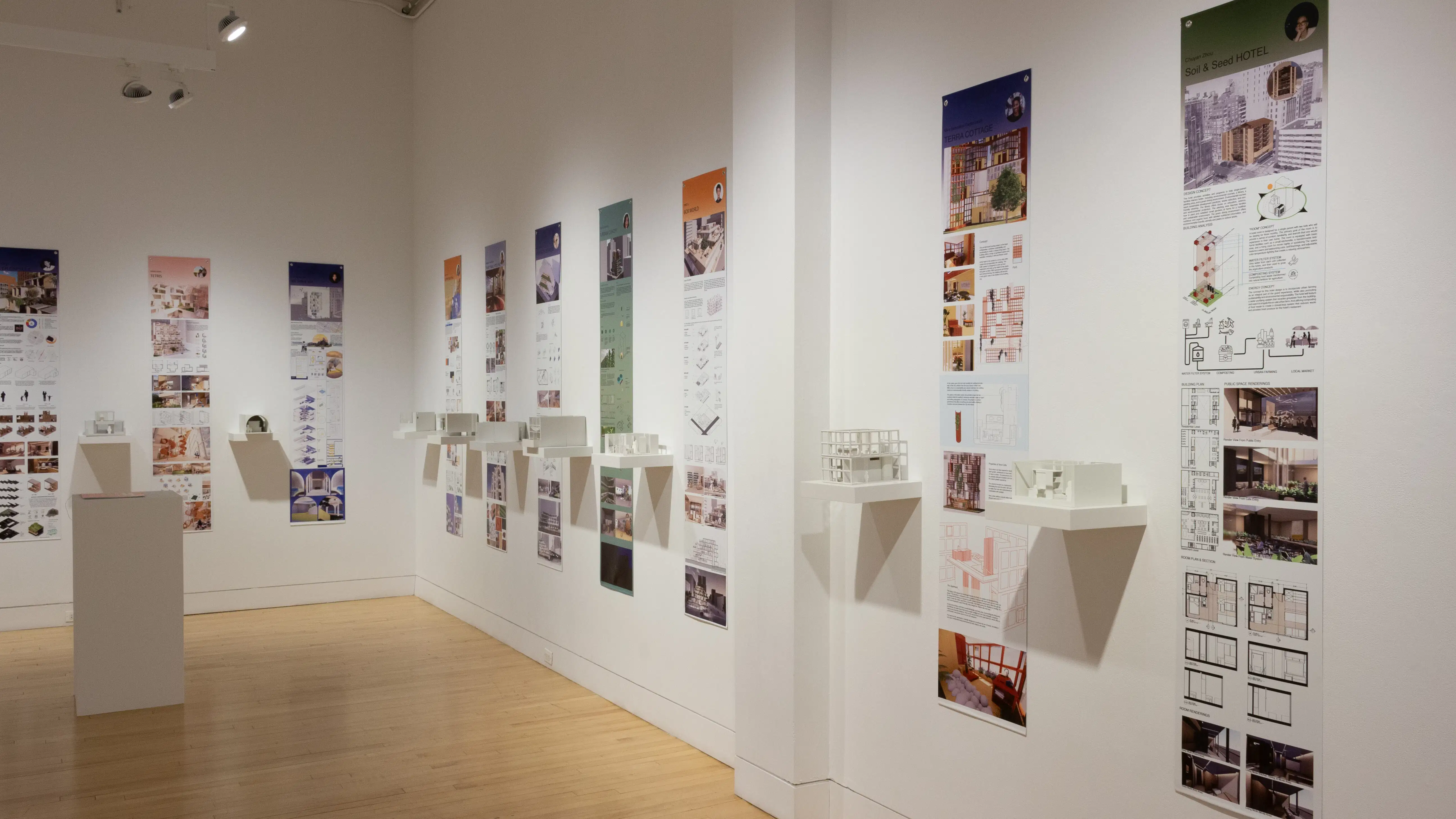 Installation image showing an angled view of two gallery walls with eight building models on shelves, and information banners for each to the right. There is a pedestal on the floor with a book on top.