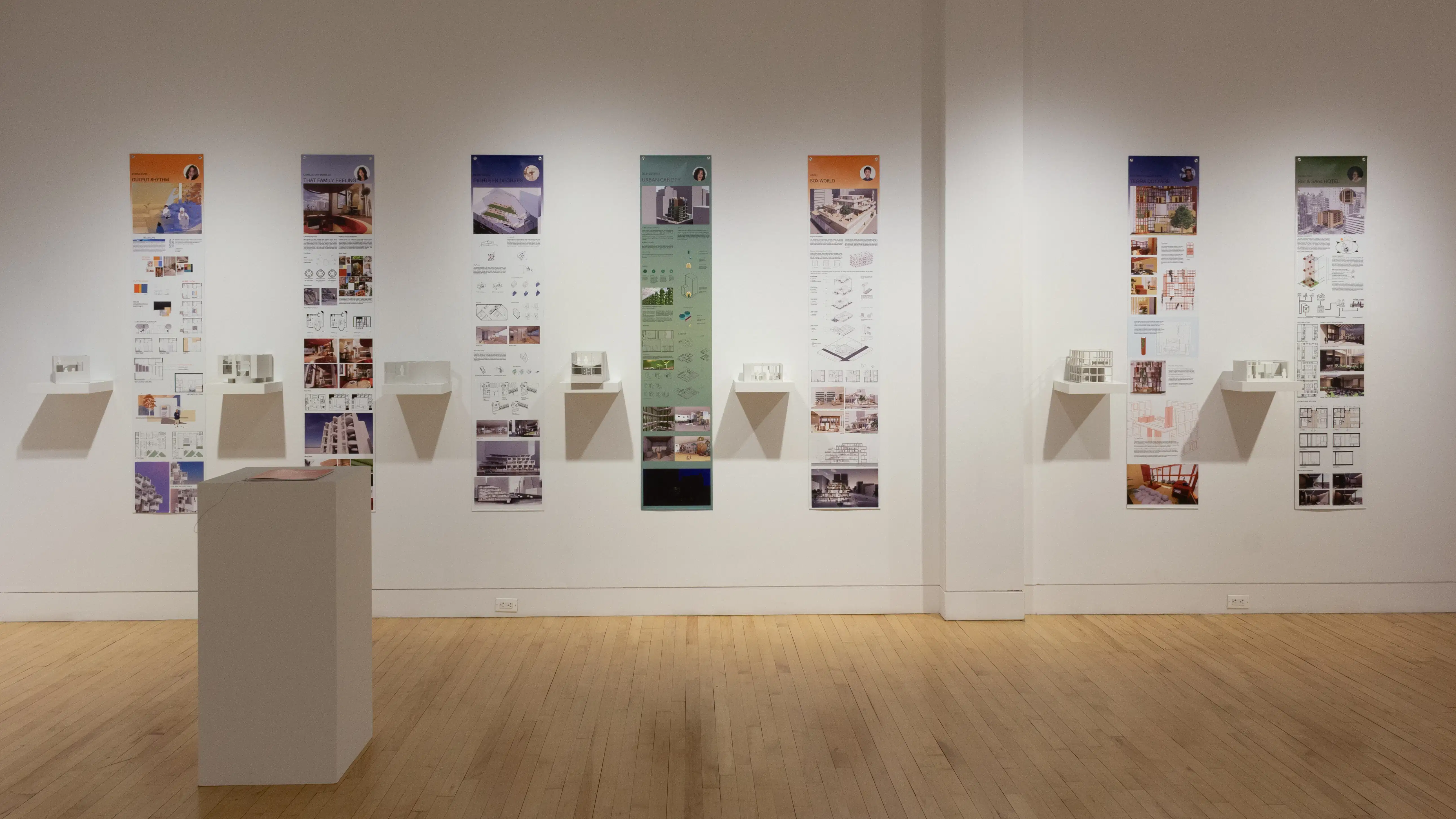 Installation image showing a straight view of a gallery wall with seven building models on shelves, and information banners for each to the right. There is a pedestal on the floor with a book on top.