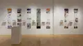 Installation image showing a straight view of a gallery wall with seven building models on shelves, and information banners for each to the right. There is a pedestal on the floor with a book on top.