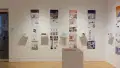 Installation image showing a straight view of two gallery walls with five building models on shelves in total, and information banners for each to the right. There is a pedestal on the floor with a book on top.