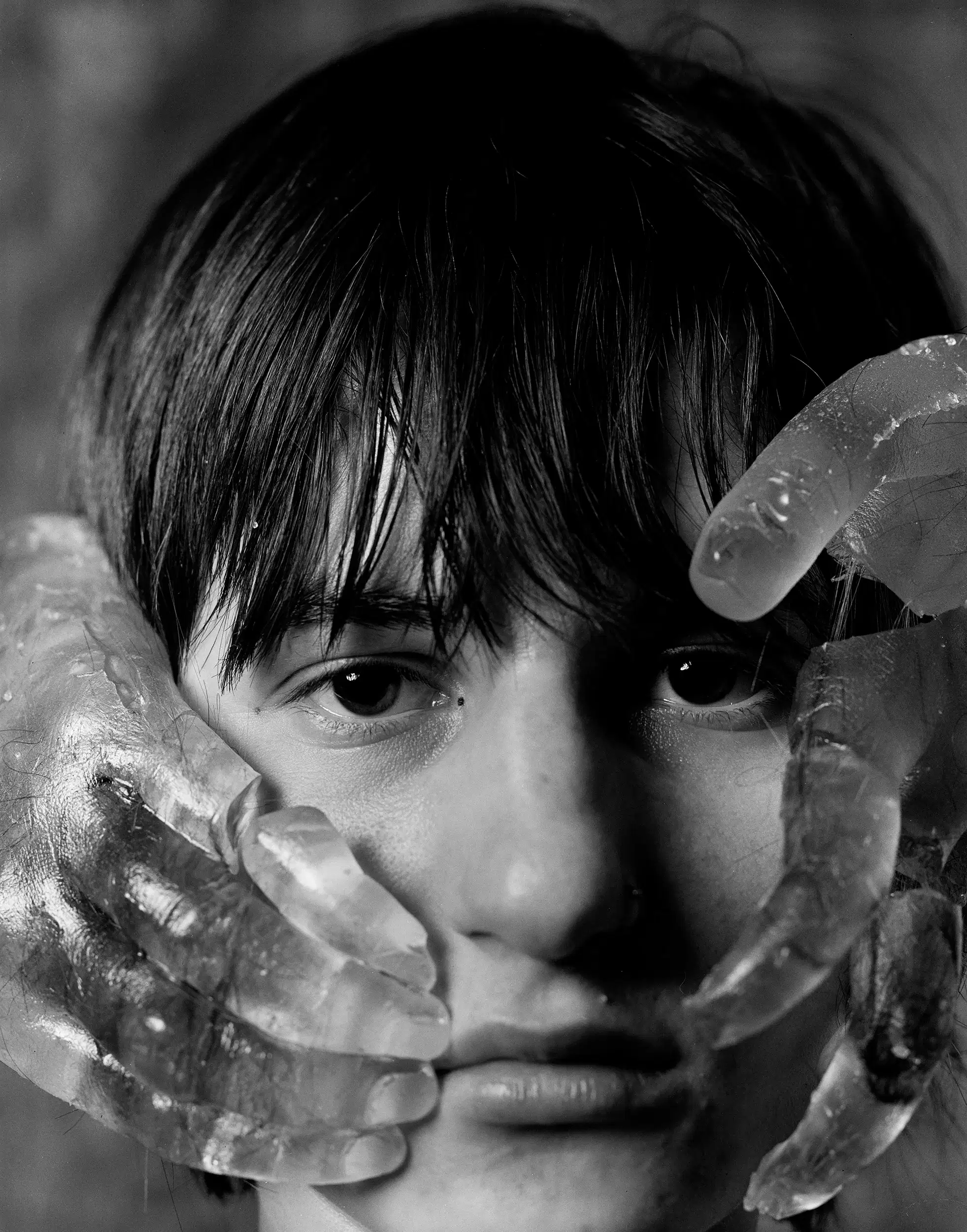 Black and white photo of a person's face obscured by translucent, ice-like material, with hands raised to their head