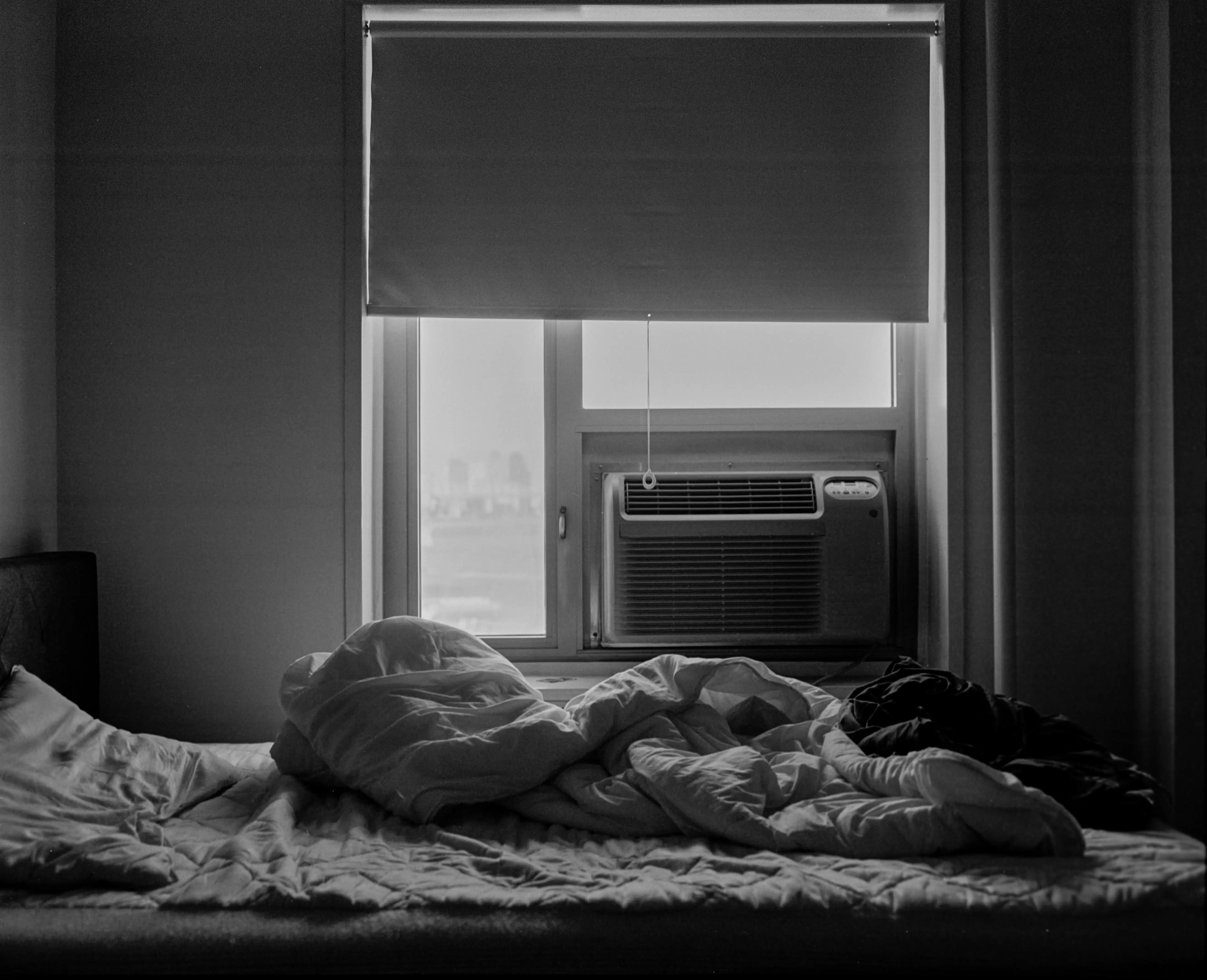 The black and white photo shows an unmade bed and a window air conditioner under half-drawn curtains